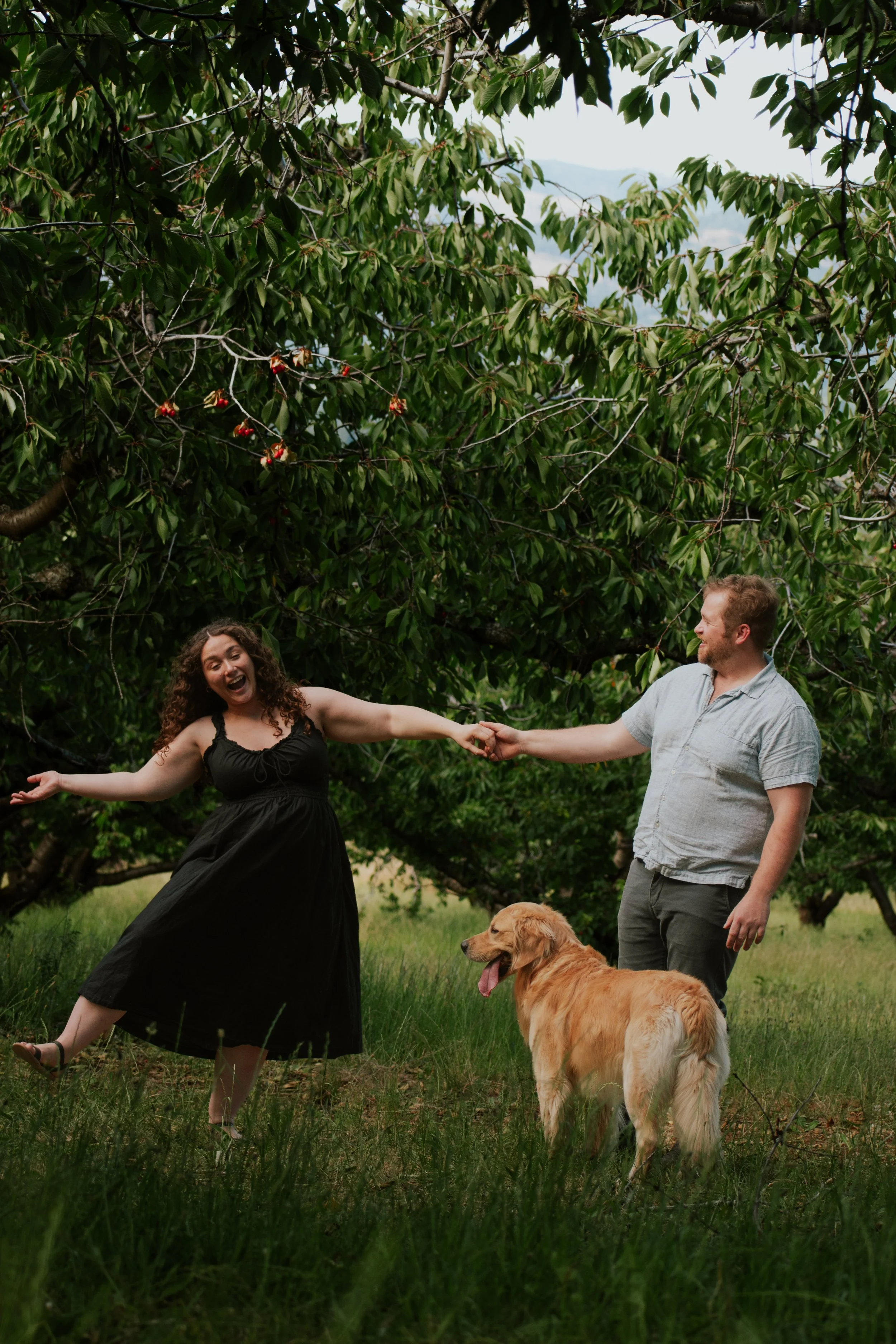 A woman in a black dress dancing joyfully outdoors with a man holding her hand, a golden retriever dog standing nearby, and lush green trees in the background.