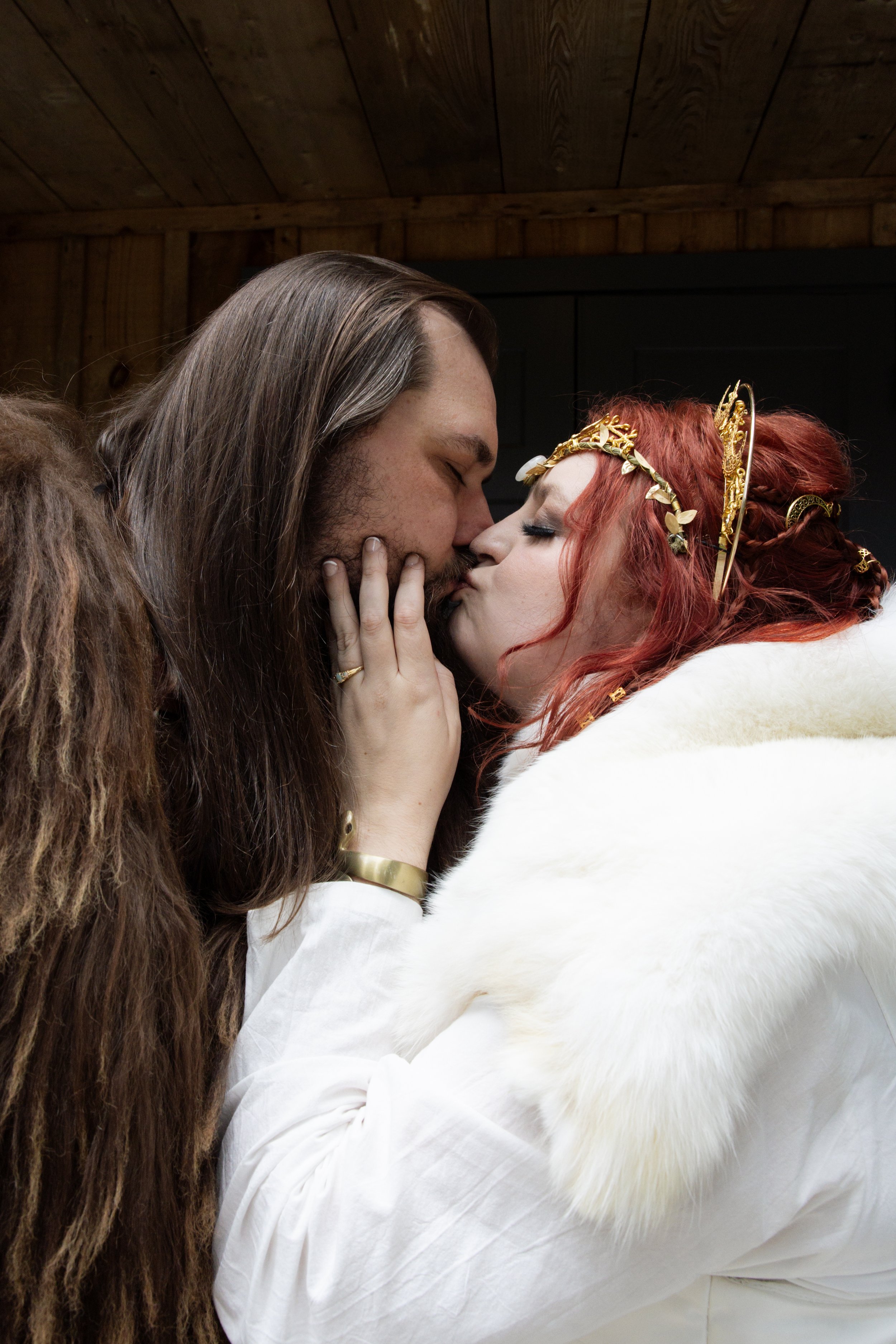 A man and a woman with fantasy makeup and accessories touch foreheads and close eyes in a warm intimate moment indoors, with wooden ceiling and dark wall background.