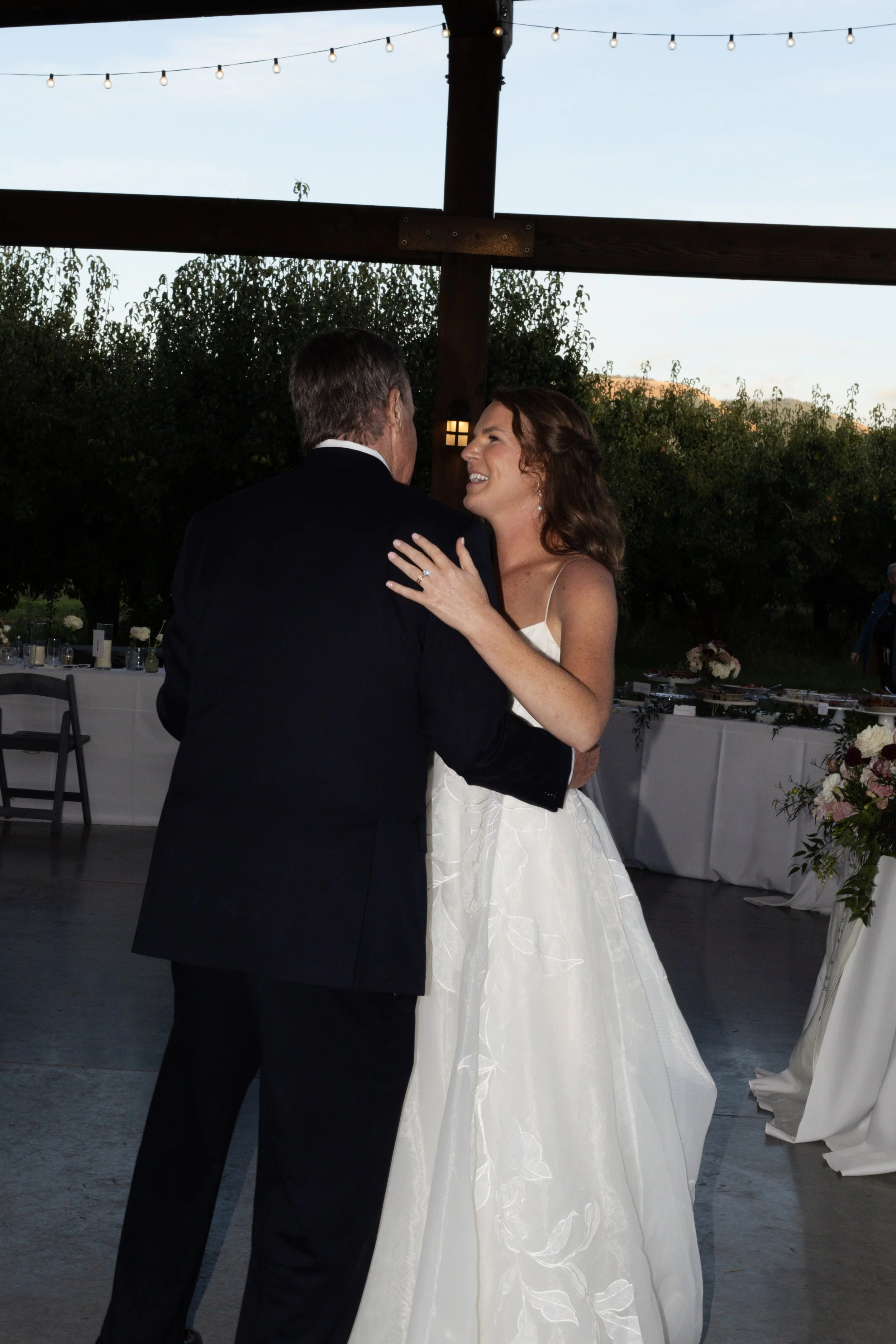A bride in a white wedding dress dancing with a groom in a black suit at an outdoor wedding reception during the evening. String lights decorate the background, and tables with floral arrangements are visible.