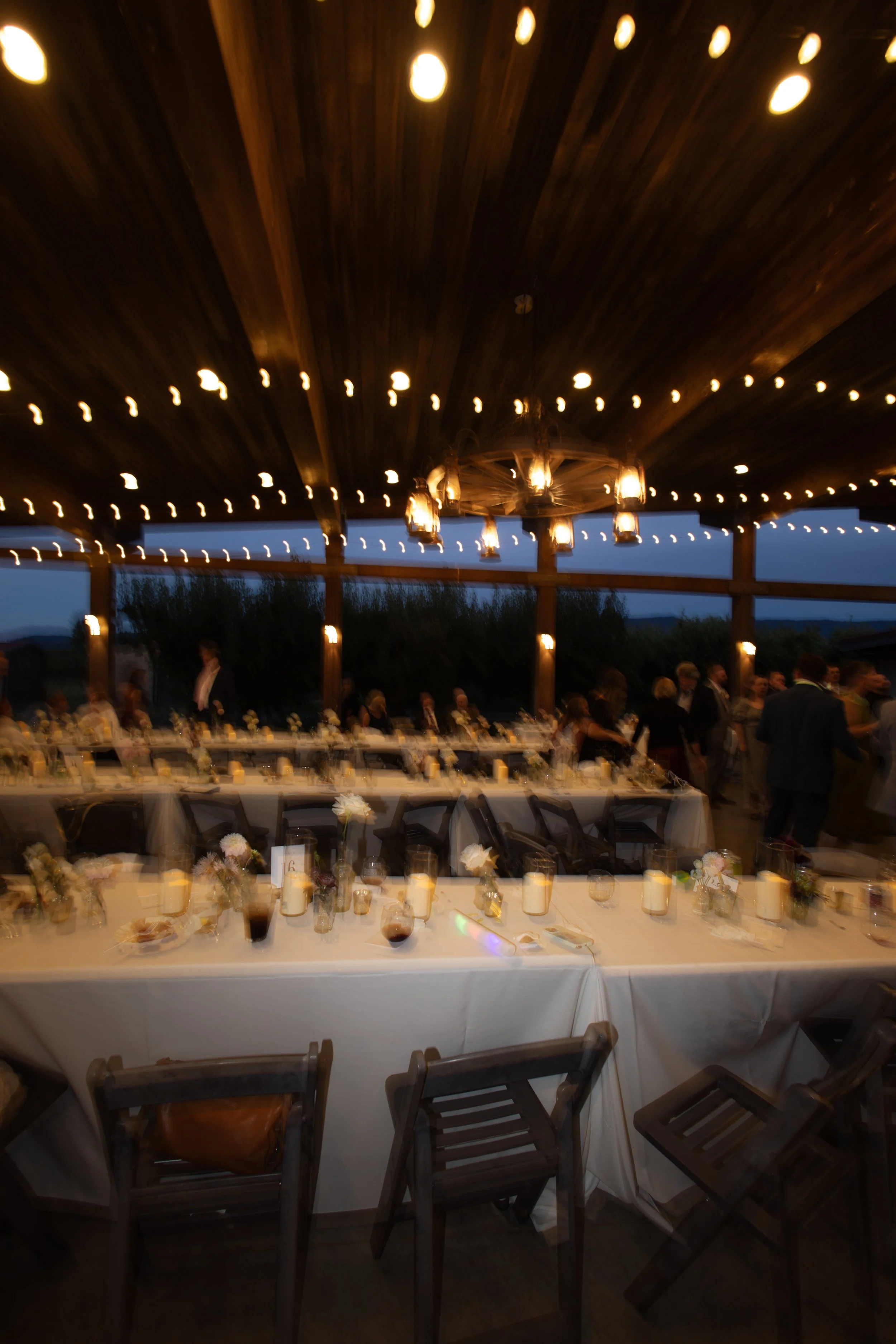 Indoor wedding reception area with long tables decorated with candles and flowers, and wooden chairs. The ceiling is decorated with string lights and a chandelier, with large windows showing an outdoor view during the evening.