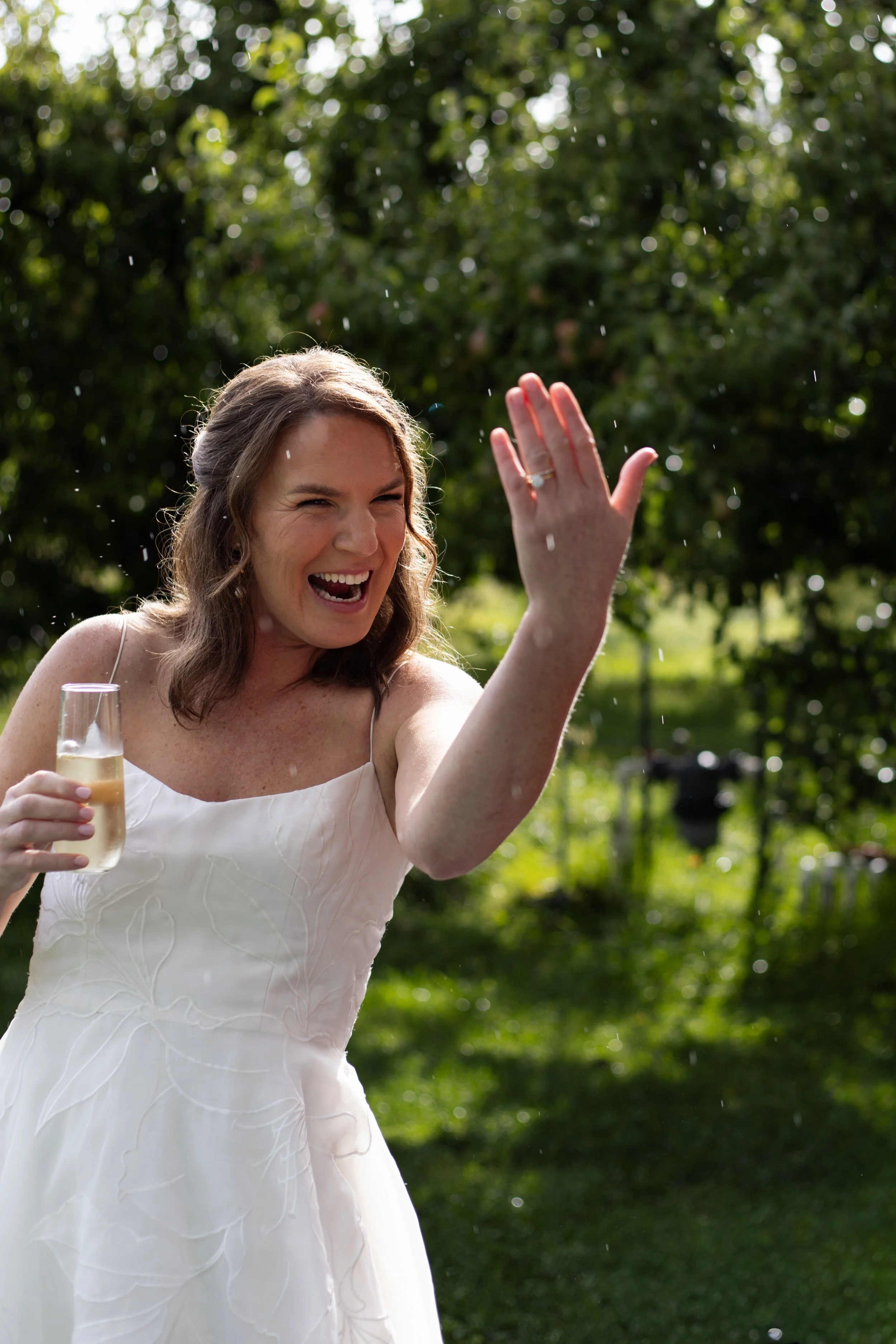 A bride smiling and holding up her wedding ring, holding a glass of champagne, with water droplets in the air and trees in the background at her wedding at Grateful Vineyards 