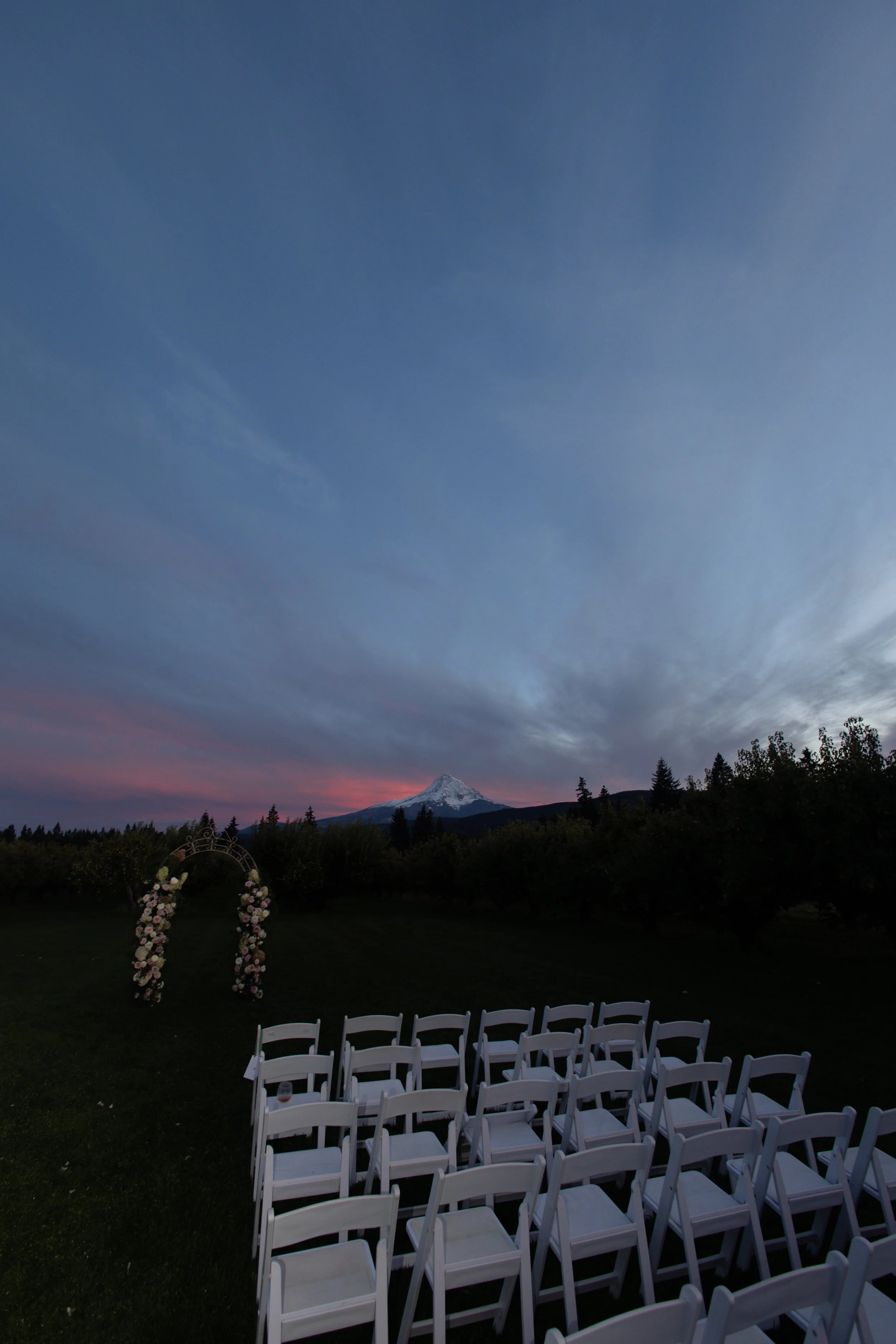 Outdoor wedding setup with white chairs arranged on a grassy field, a floral arch at the altar, and Mount Hood in the background, under a cloudy evening sky.