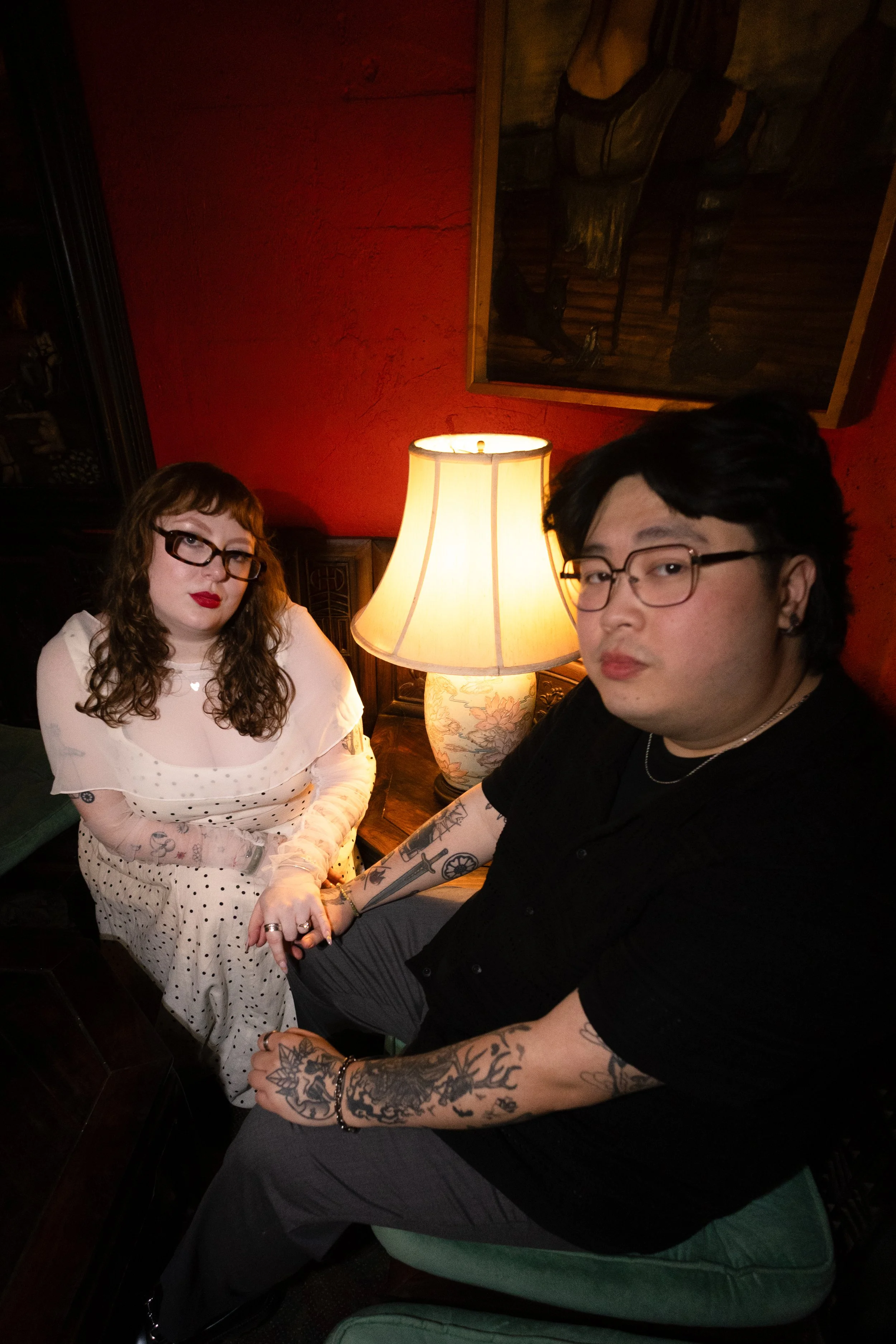 A woman with curly hair, wearing glasses and a white polka dot dress, sitting and holding hands with a man with dark hair, glasses, and tattoos on his arms, in a dimly lit room with warm lighting, red walls, a lamp, and framed artwork.