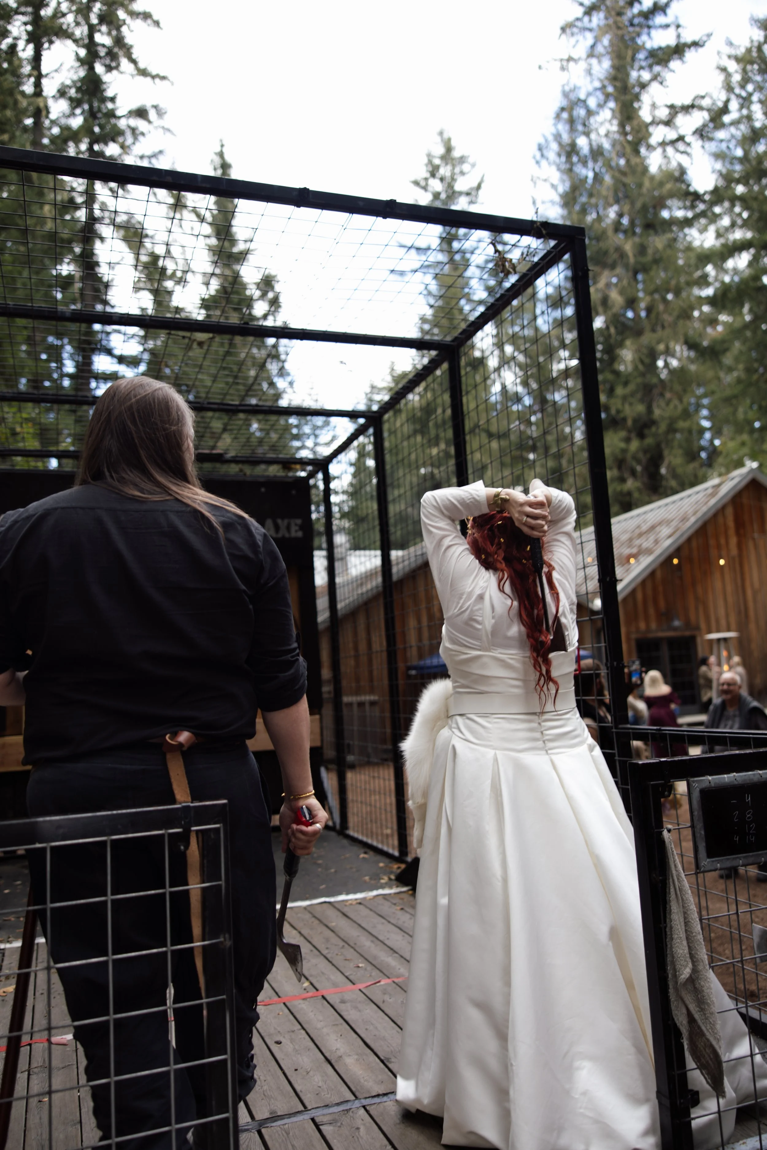 A woman with red hair in a white dress stands inside a metal cage at an outdoor event, while another woman, with brown hair, stands nearby holding a leash. There are many people in the background, and trees and a wooden building are visible.