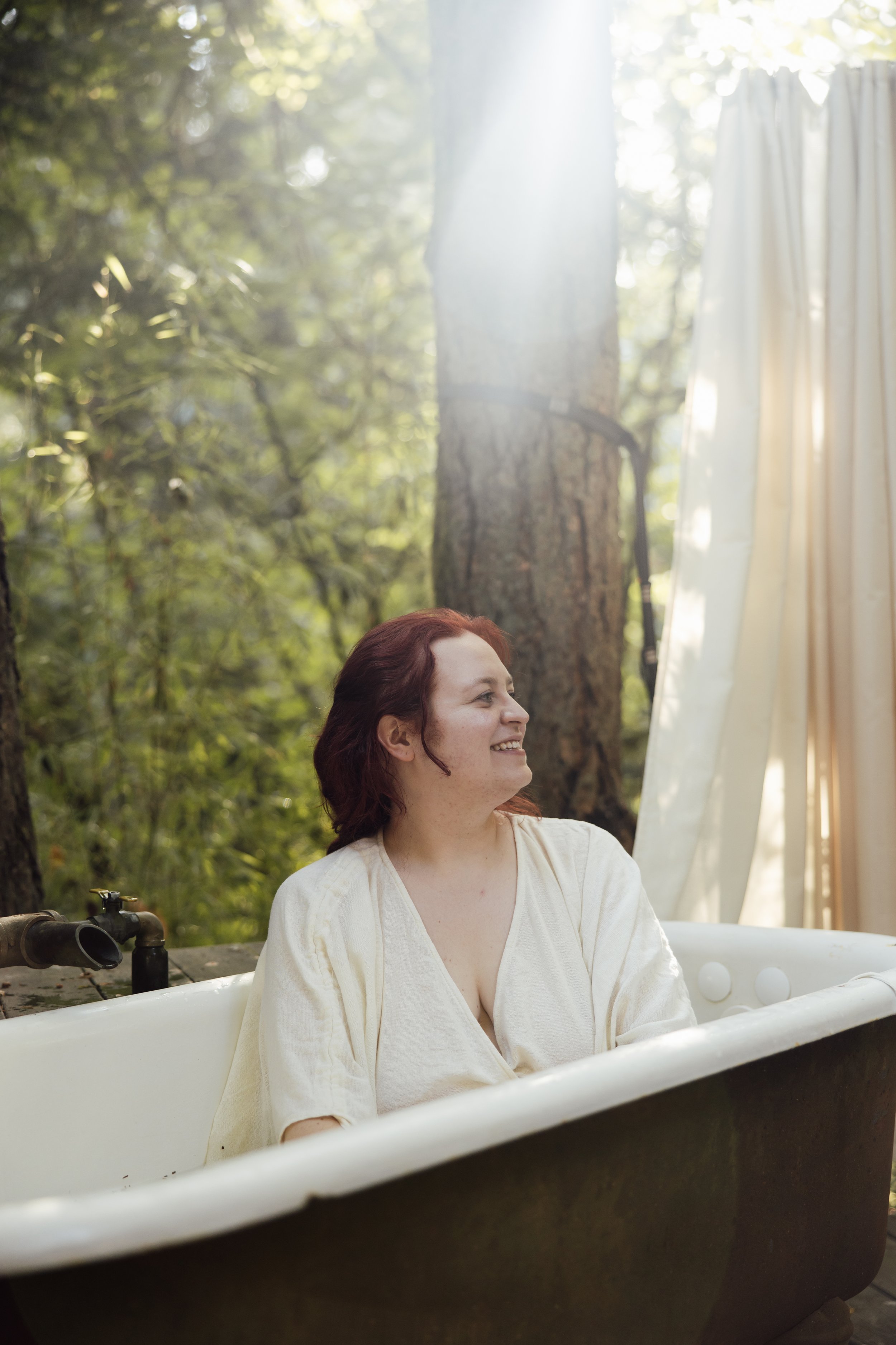 A woman with red hair smiling while sitting in a white bathtub outdoors in a forest, with sunlight filtering through trees and a curtain in the background.