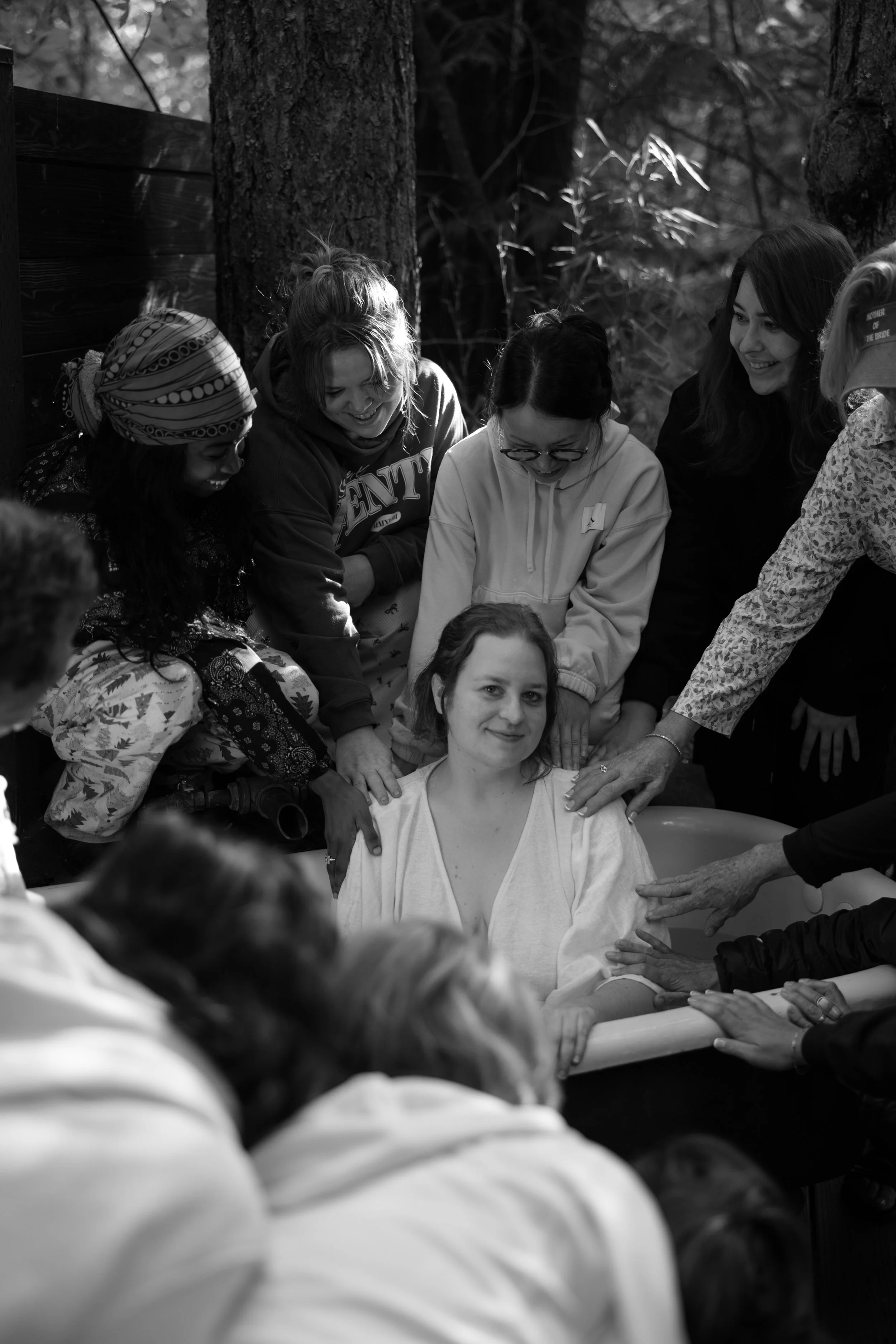 Group of women gathered around a woman sitting in a bathtub outdoors, participating in a baptism ceremony in a wooded area.