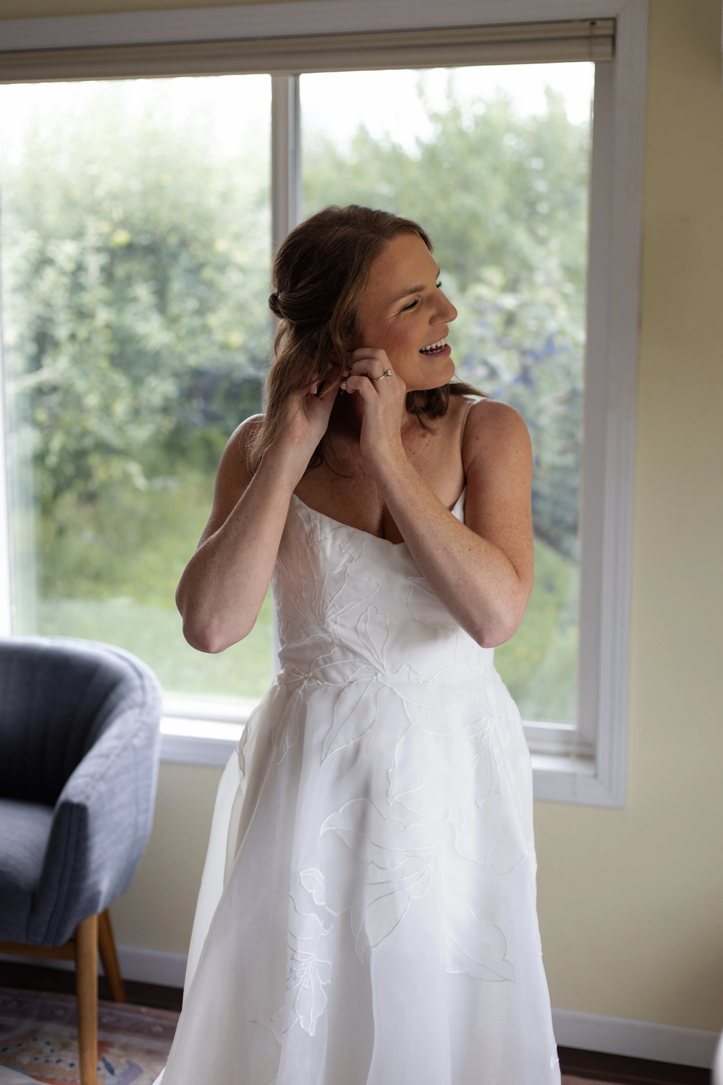 A woman in a white dress smiling while putting on earrings inside a room with a large window showing trees outside.