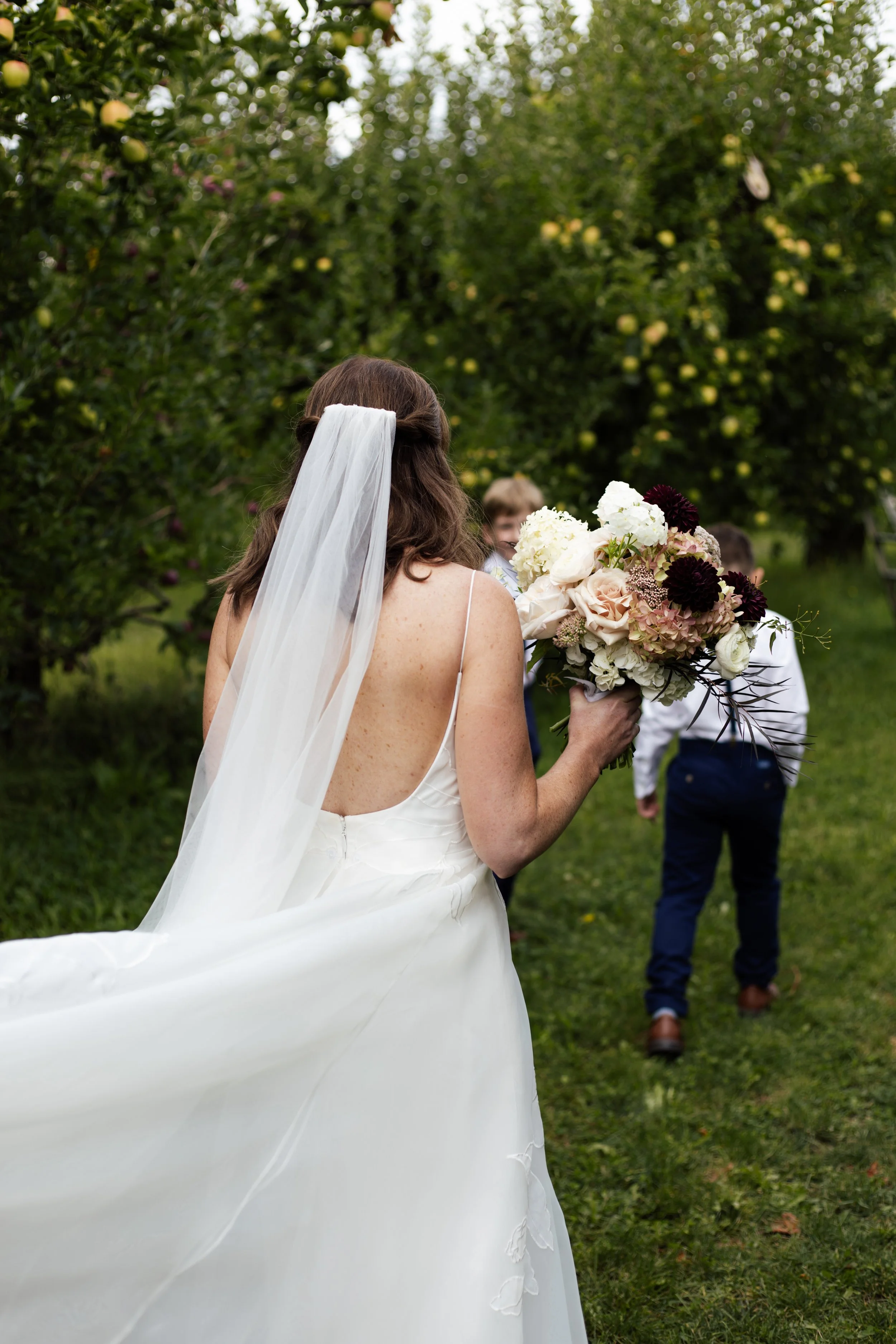 A bride with a veil and a white wedding dress holding a large bouquet of flowers, looking at a group of people in an orchard with trees bearing green apples.