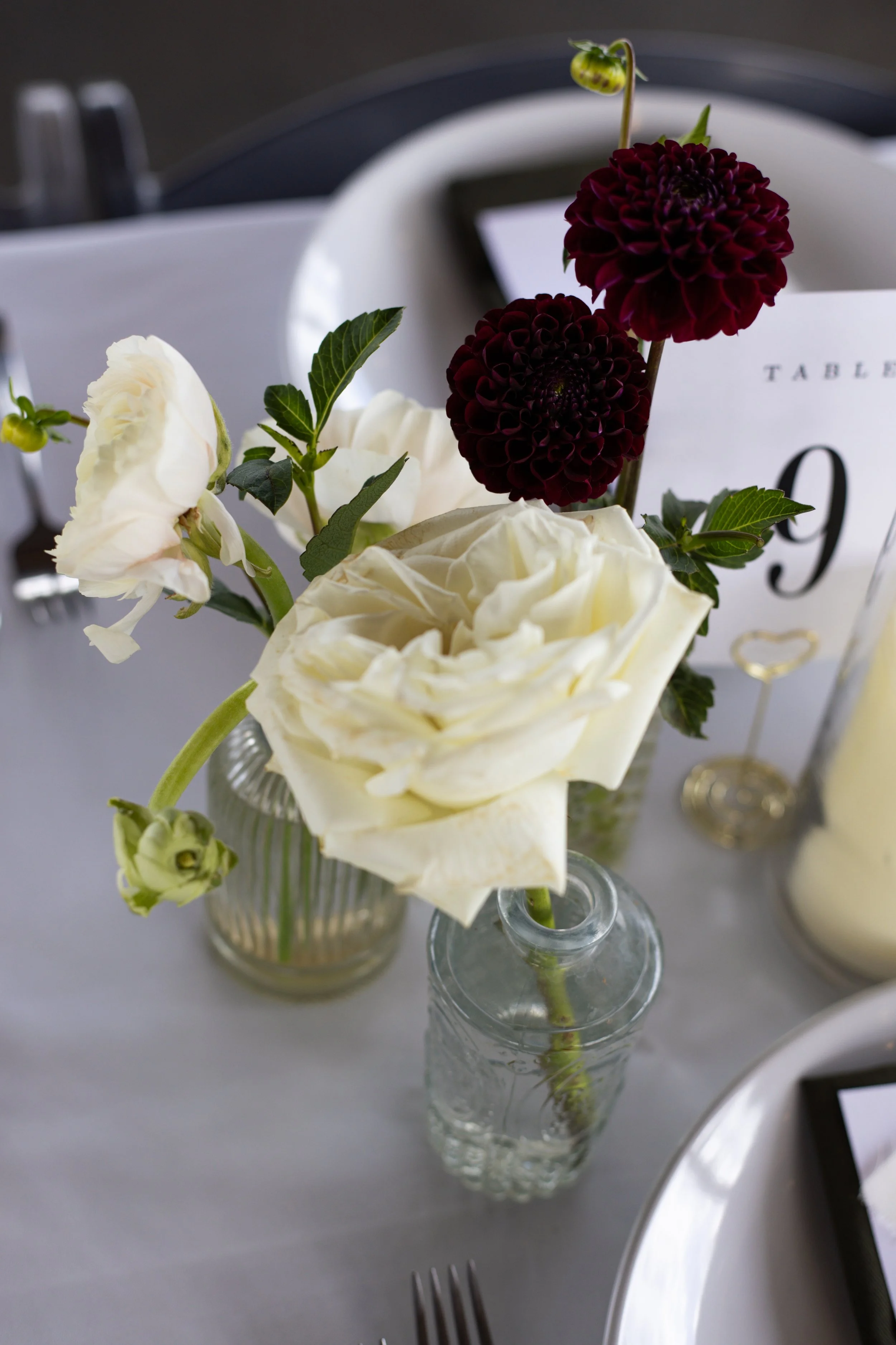 A small bouquet of white and burgundy flowers in a clear glass vase on a gray table, with a table number card visible in the background.