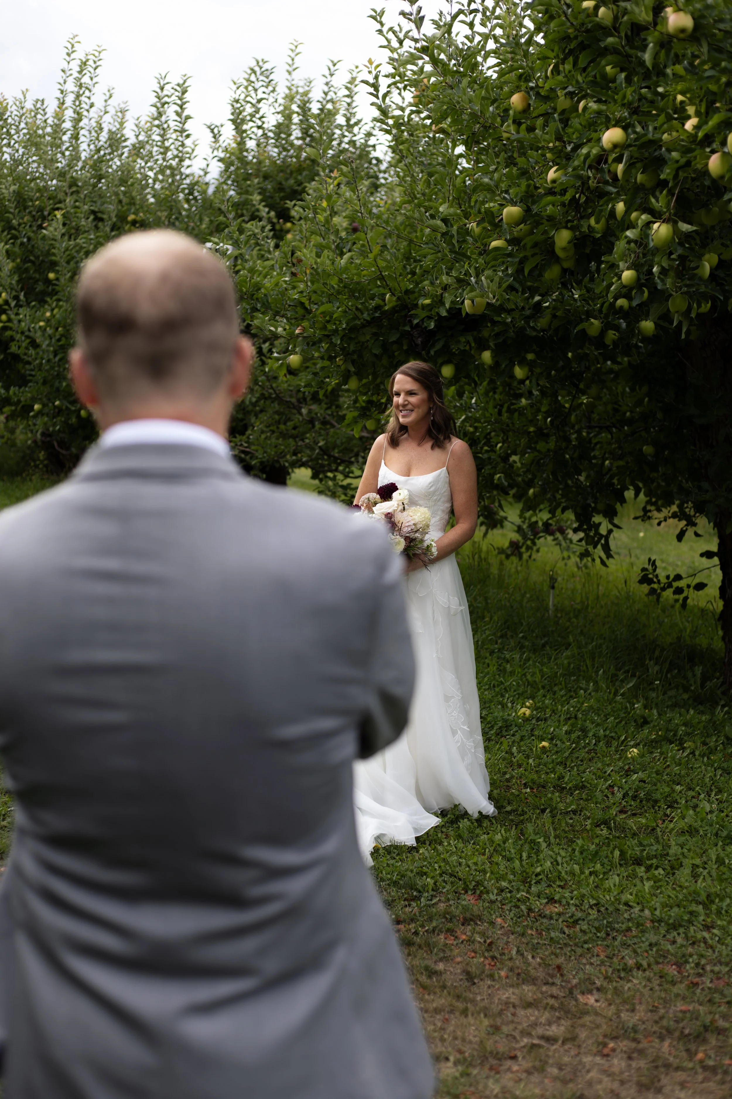 A bride in a white wedding dress smiling at a groom during an outdoor wedding ceremony in an apple orchard, with green trees and apples in the background.