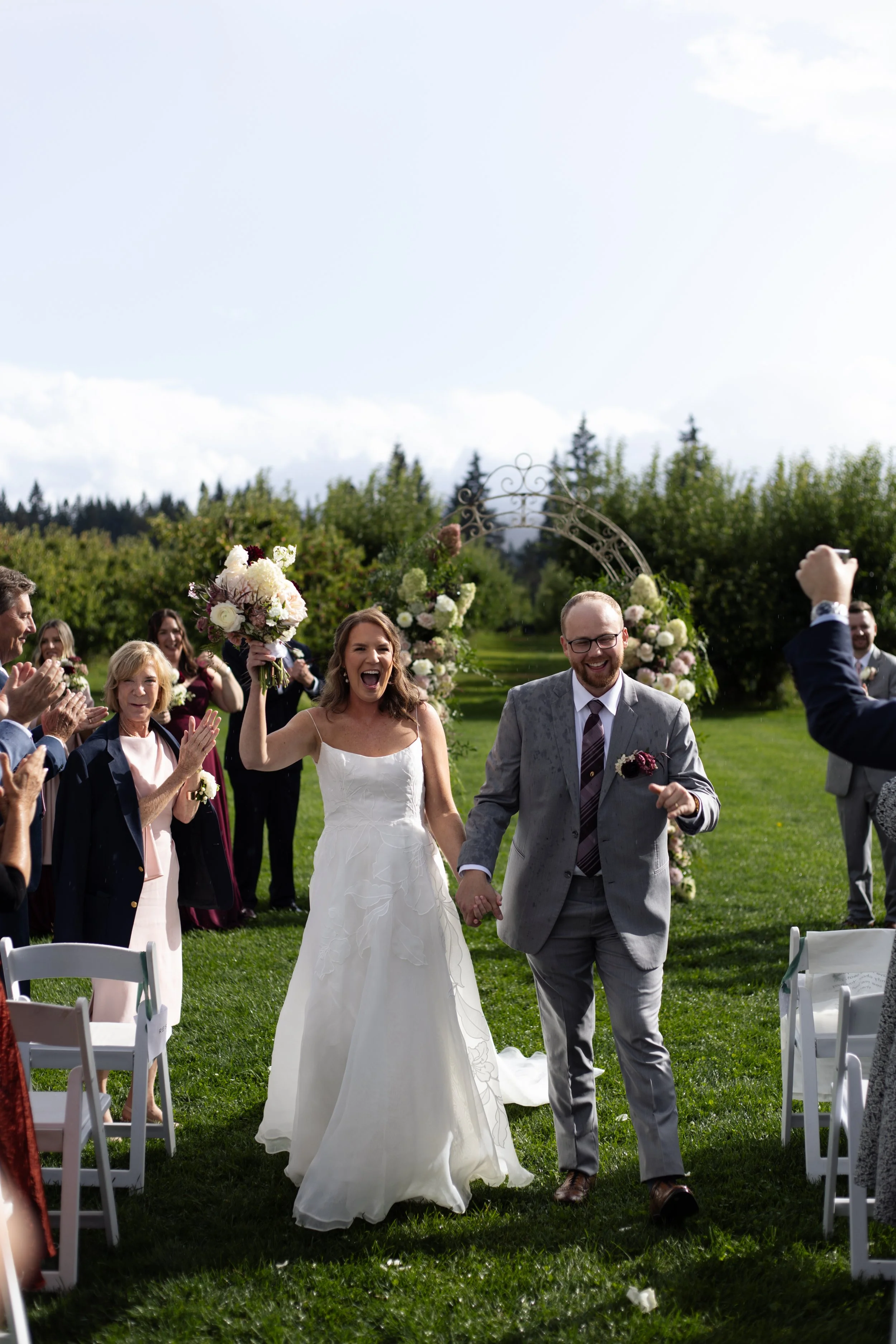 A newlywed couple celebrates their wedding outdoors with smiling guests around, holding bouquets, with chairs and a floral arch in the background.