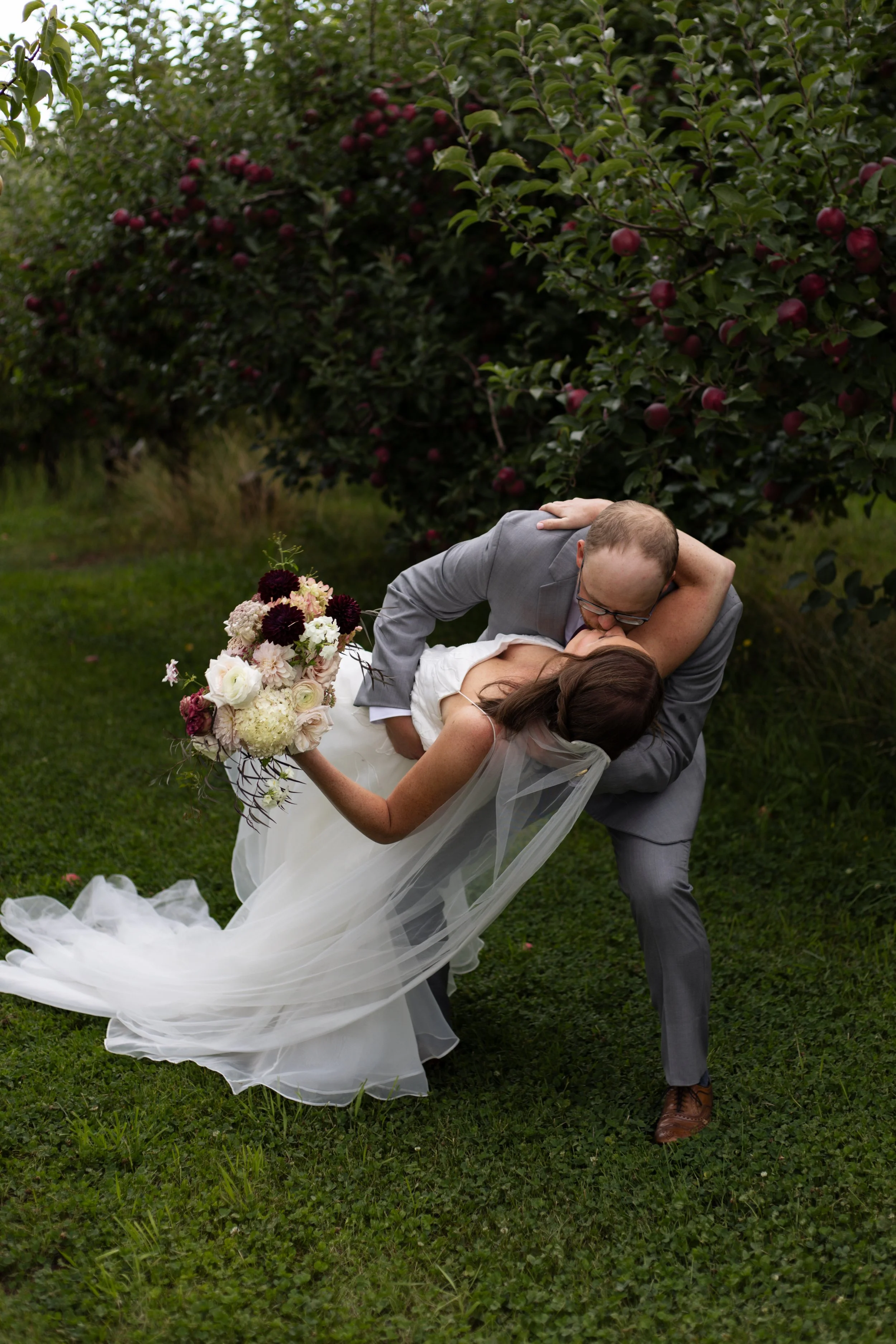 A groom lifts and kisses a bride outdoors in a garden with apple trees, while the bride holds a large bouquet of flowers and wears a white wedding dress with a train.