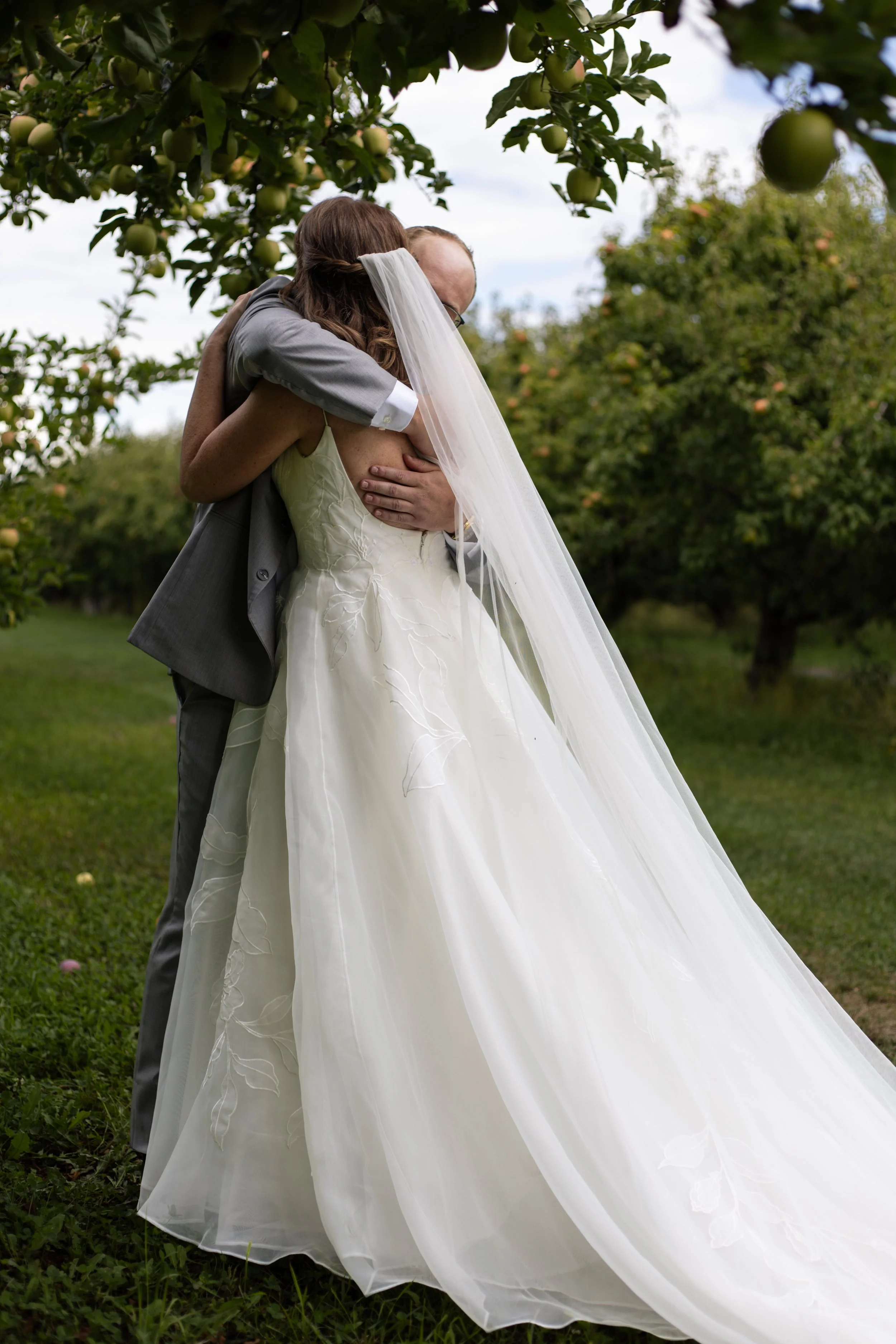 A bride and groom embrace in a garden with apple trees, with the bride wearing a white wedding dress and veil, and the groom dressed in a gray suit.