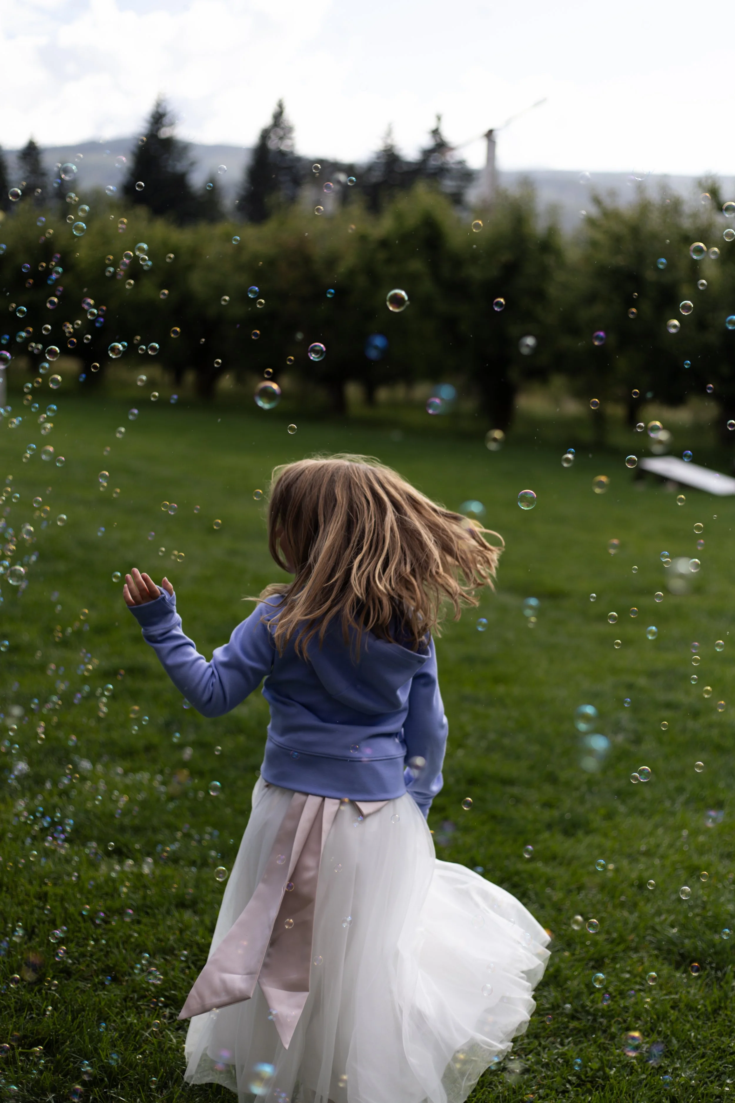 A young girl with long blonde hair is outside on grass, wearing a white dress with a pink sash, playing with bubbles during daytime with trees and a cloudy sky in the background.