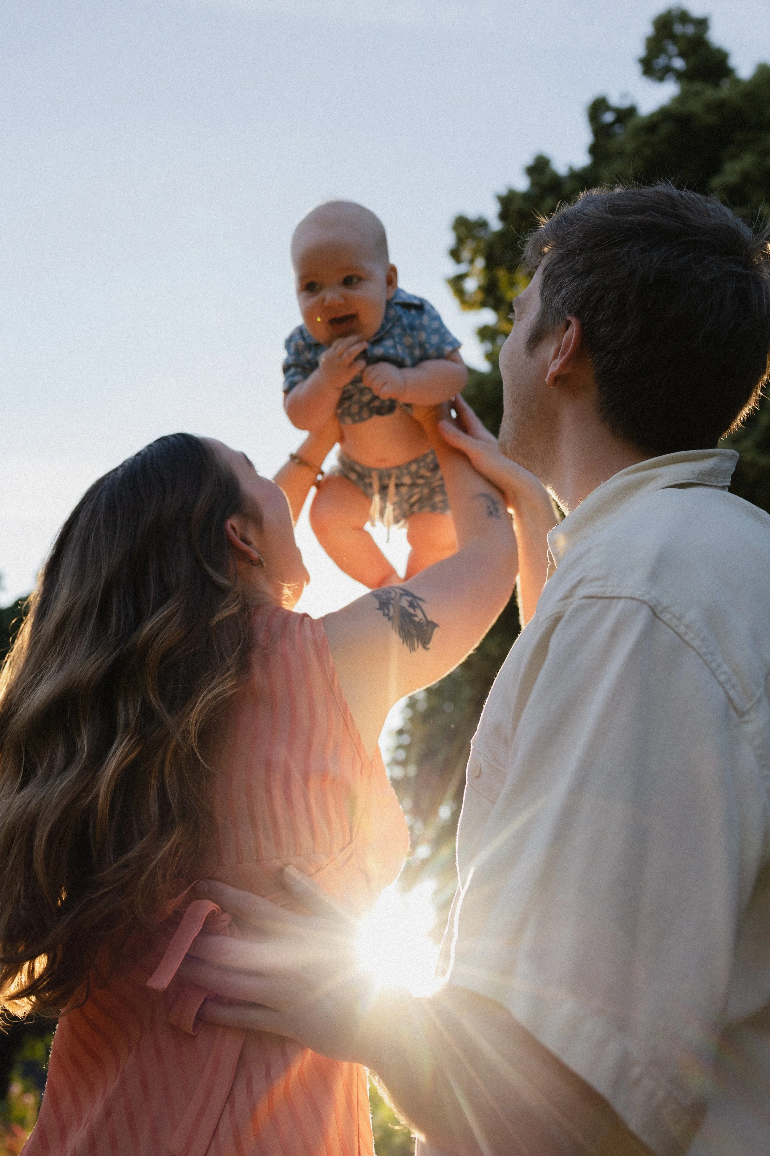 A family of three enjoying a moment outdoors at sunset, with a woman lifting a baby and a man supporting her from the other side, surrounded by trees.