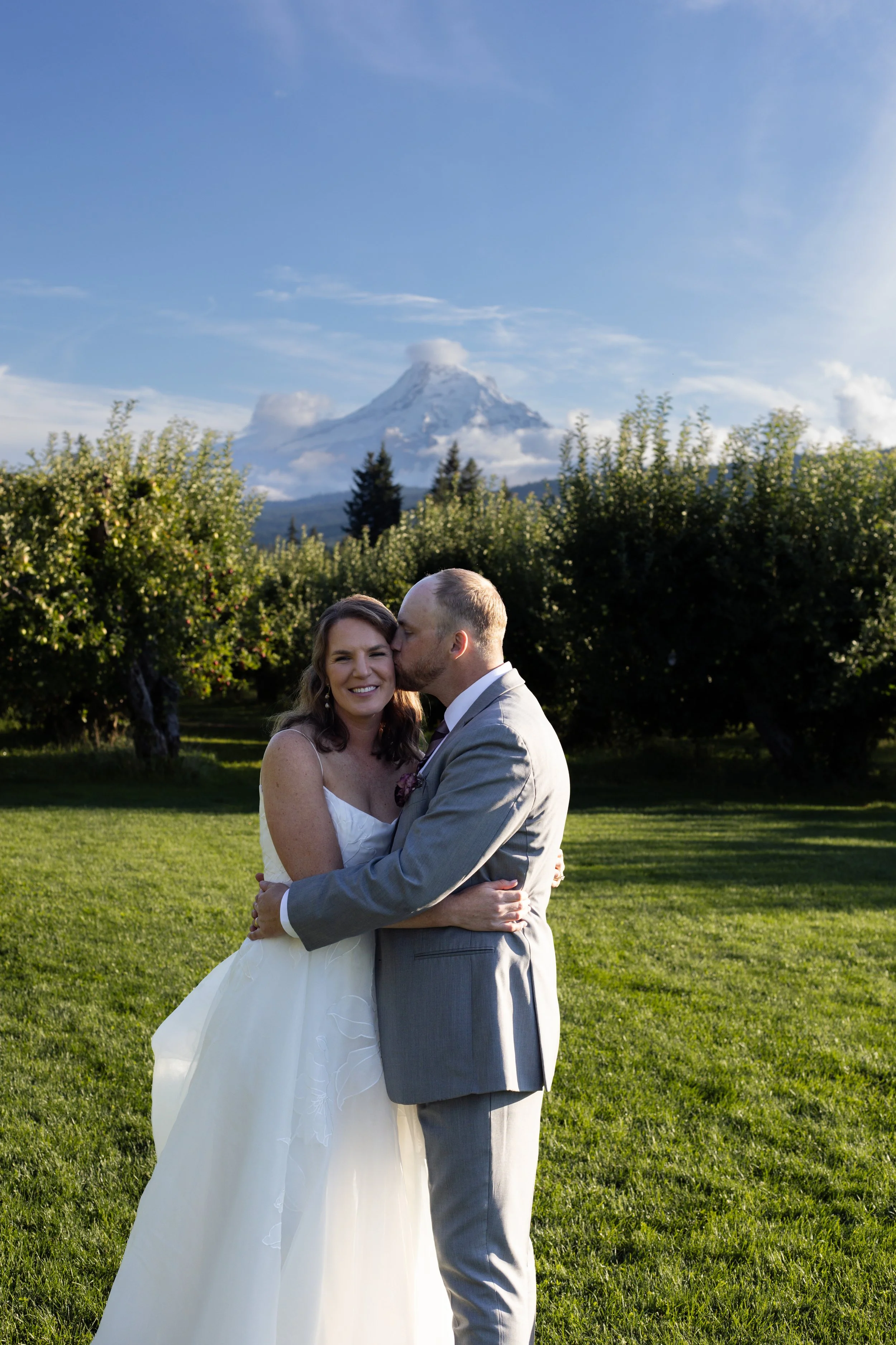 A bride and groom embrace outdoors with a snow-capped mountain and blue sky in the background.