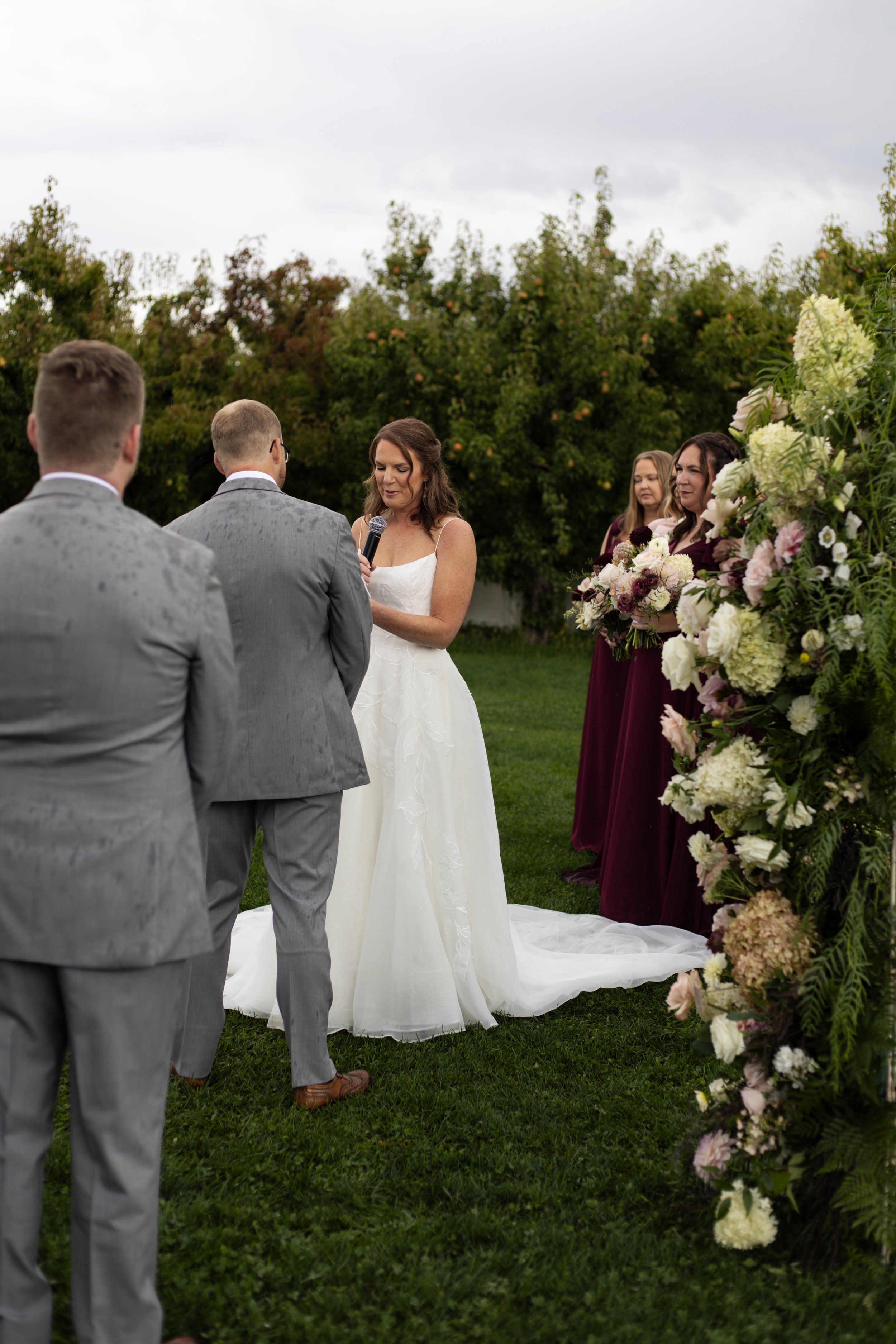 A wedding ceremony taking place outdoors with a bride holding a microphone, two grooms in gray suits facing her, and three women in burgundy dresses standing behind a floral arrangement. The scene is set on a grassy area with trees in the background 