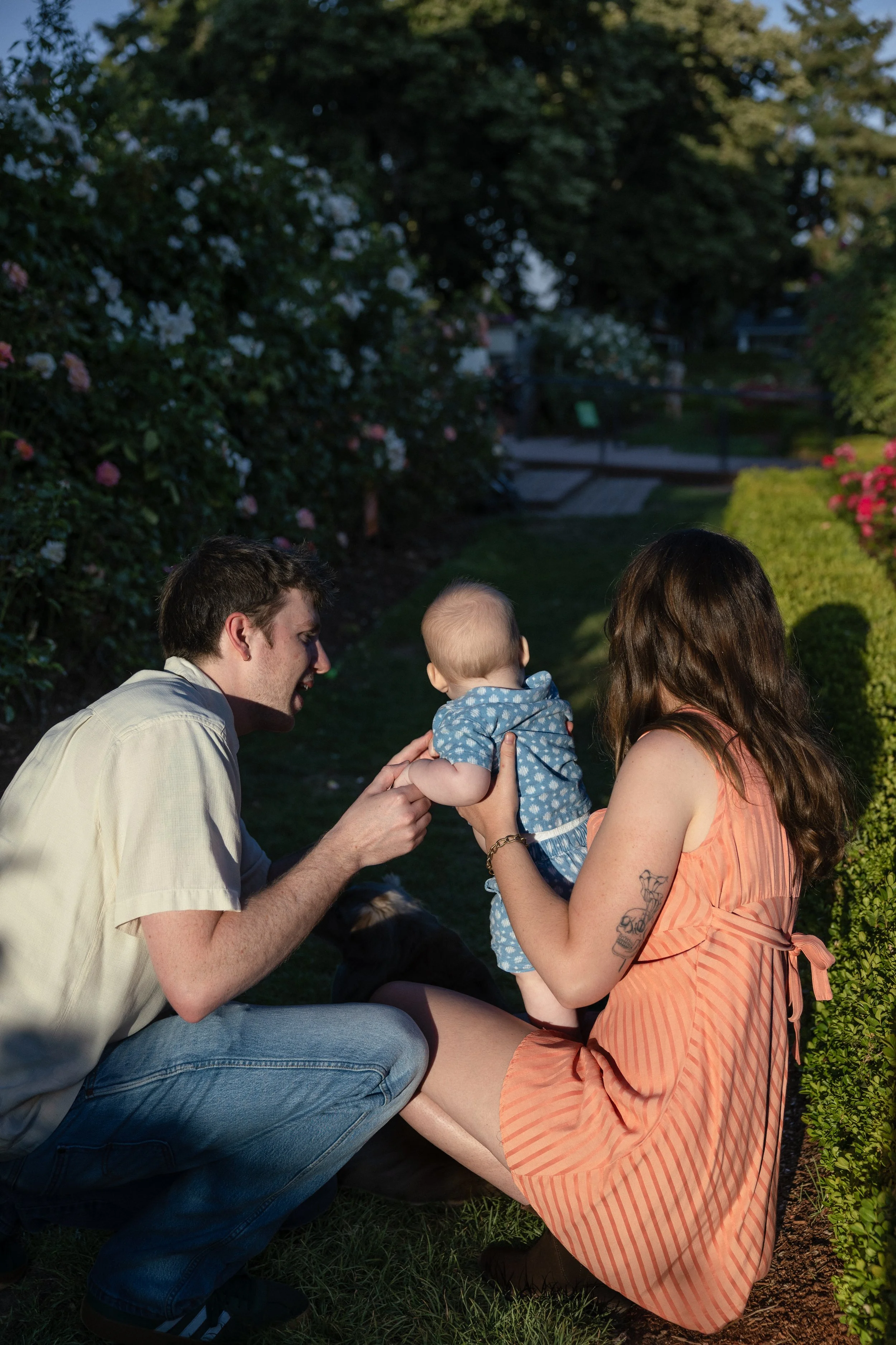 A family of three, likely parents and their child, spending time outdoors in a garden with blooming bushes and trees. The father is kneeling, holding the child's hands, while the mother is holding the child, who is dressed in a blue polka dot outfit.