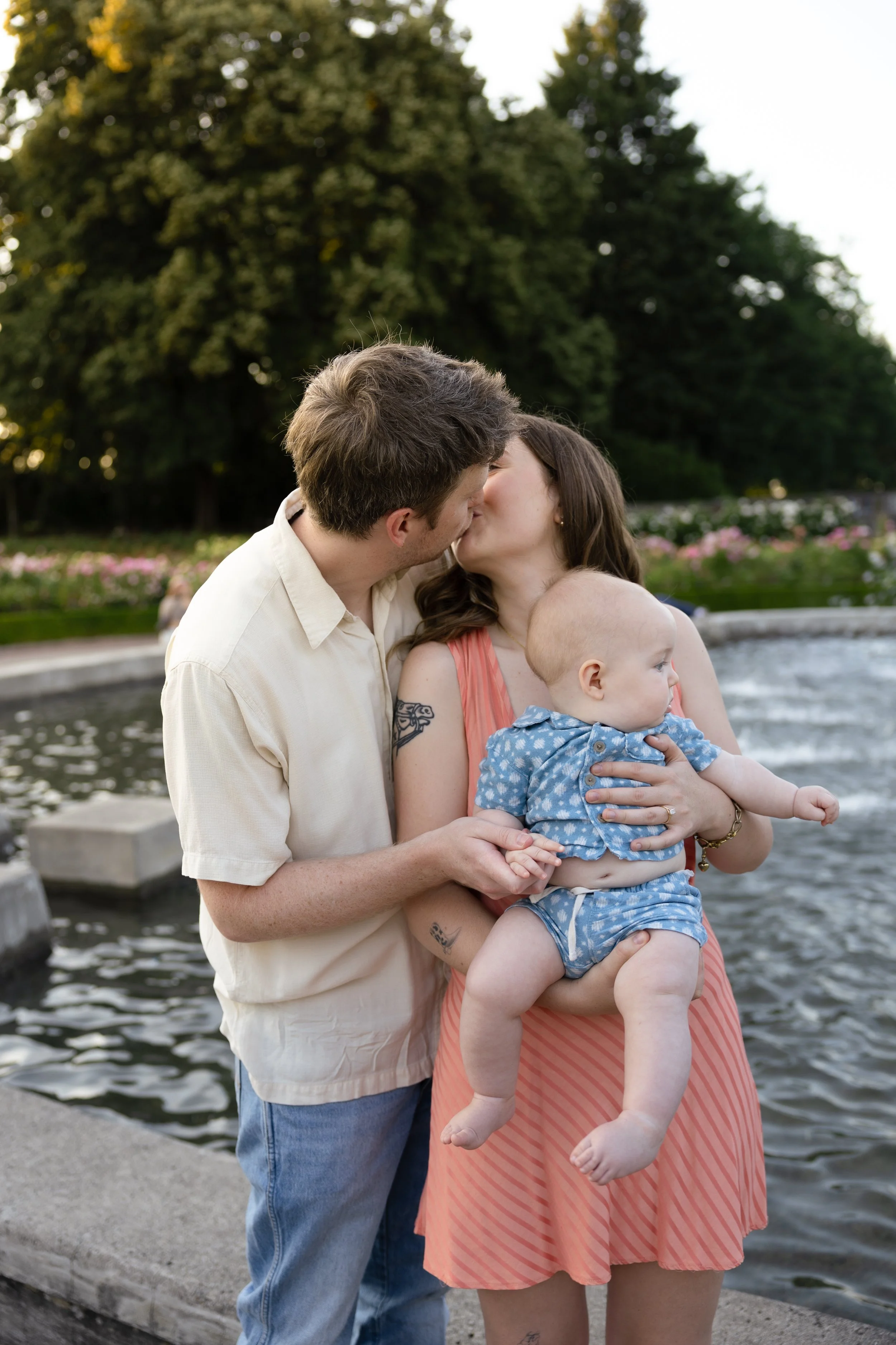 A family of three sharing a kiss outdoors by a body of water, with trees in the background.