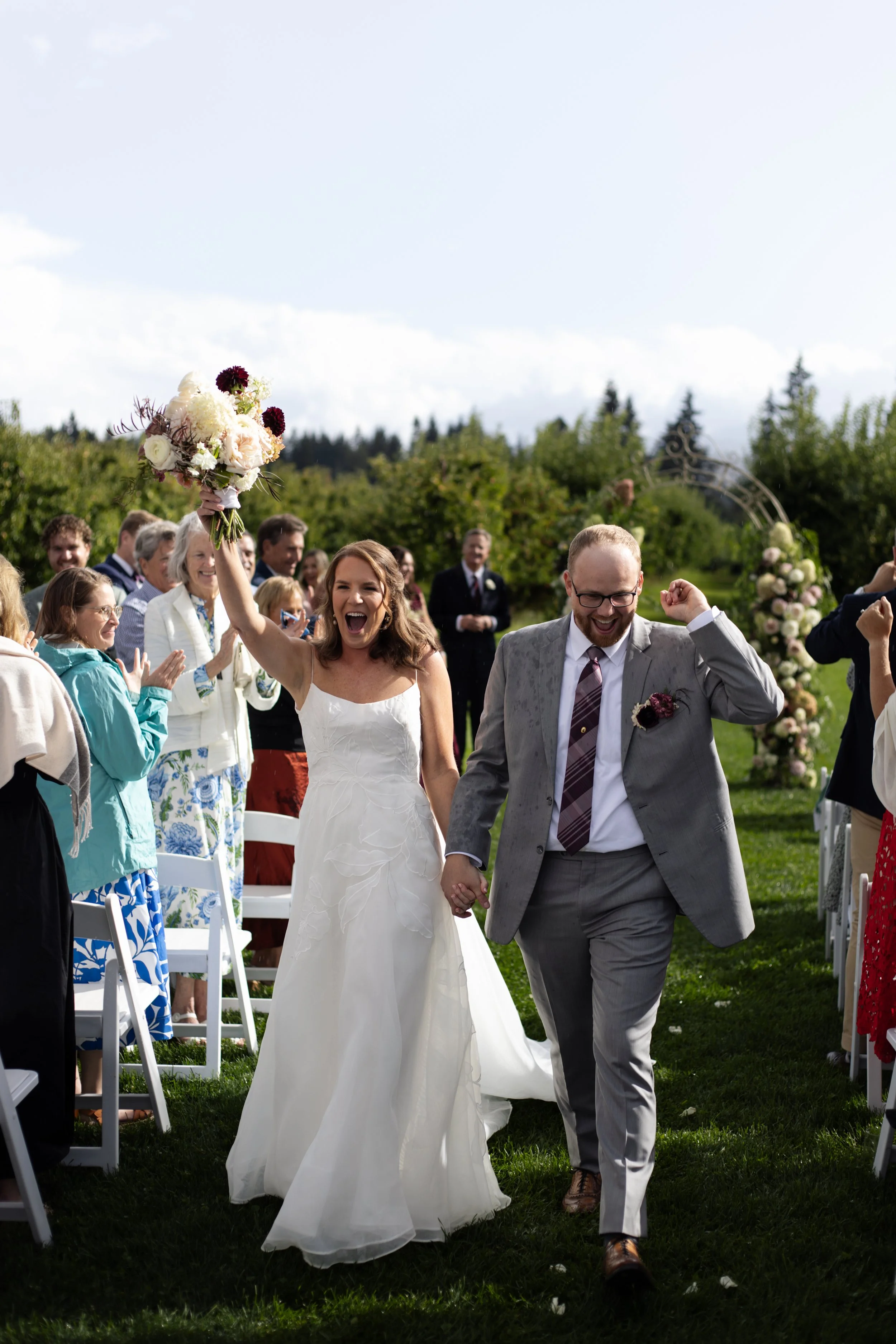 A bride and groom celebrating outdoors at their wedding, with the bride holding a bouquet and smiling, while the groom is smiling and wearing a gray suit. Guests are clapping and smiling in the background, with a floral arch in the distance.