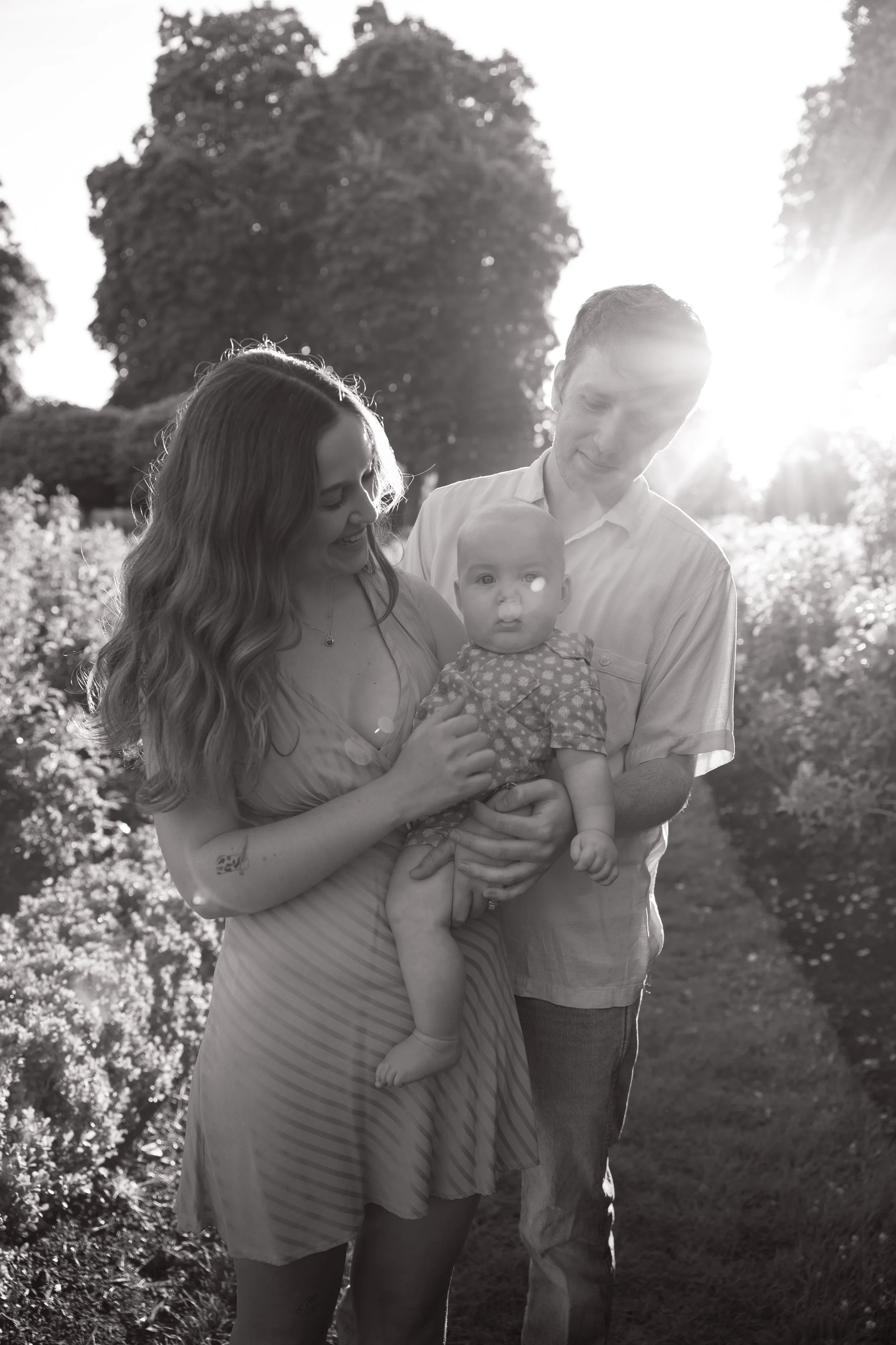 A smiling family of three, including a woman, a man, and a baby, is outdoors during the daytime with bright sunlight behind them. The woman is holding the baby, and the man is standing close, all looking down at the baby.