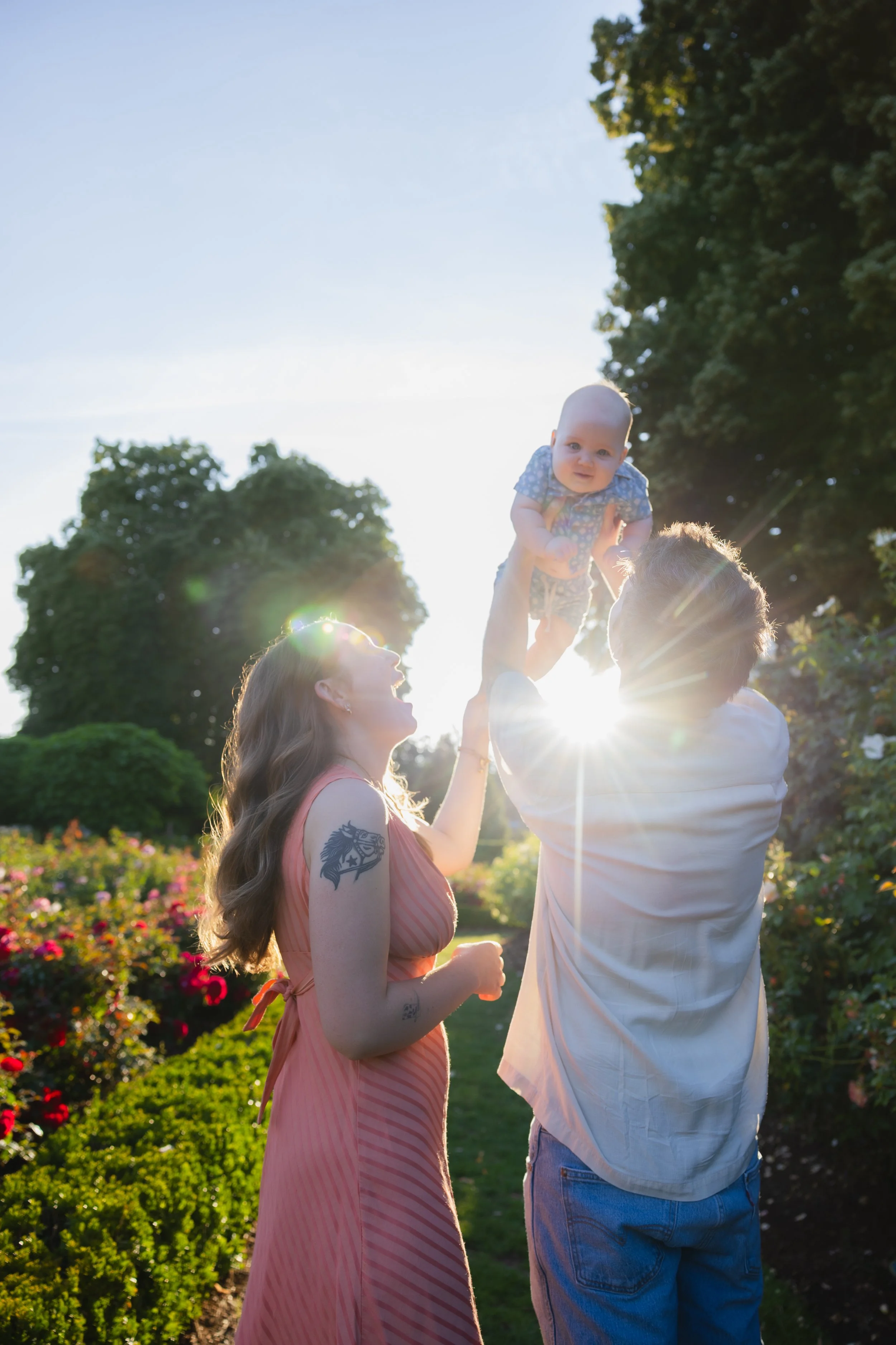 A woman with long wavy hair and tattoos on her arm lifting a baby boy with short blonde hair into the air, with a man holding him and a garden with flowers and large trees in the background.