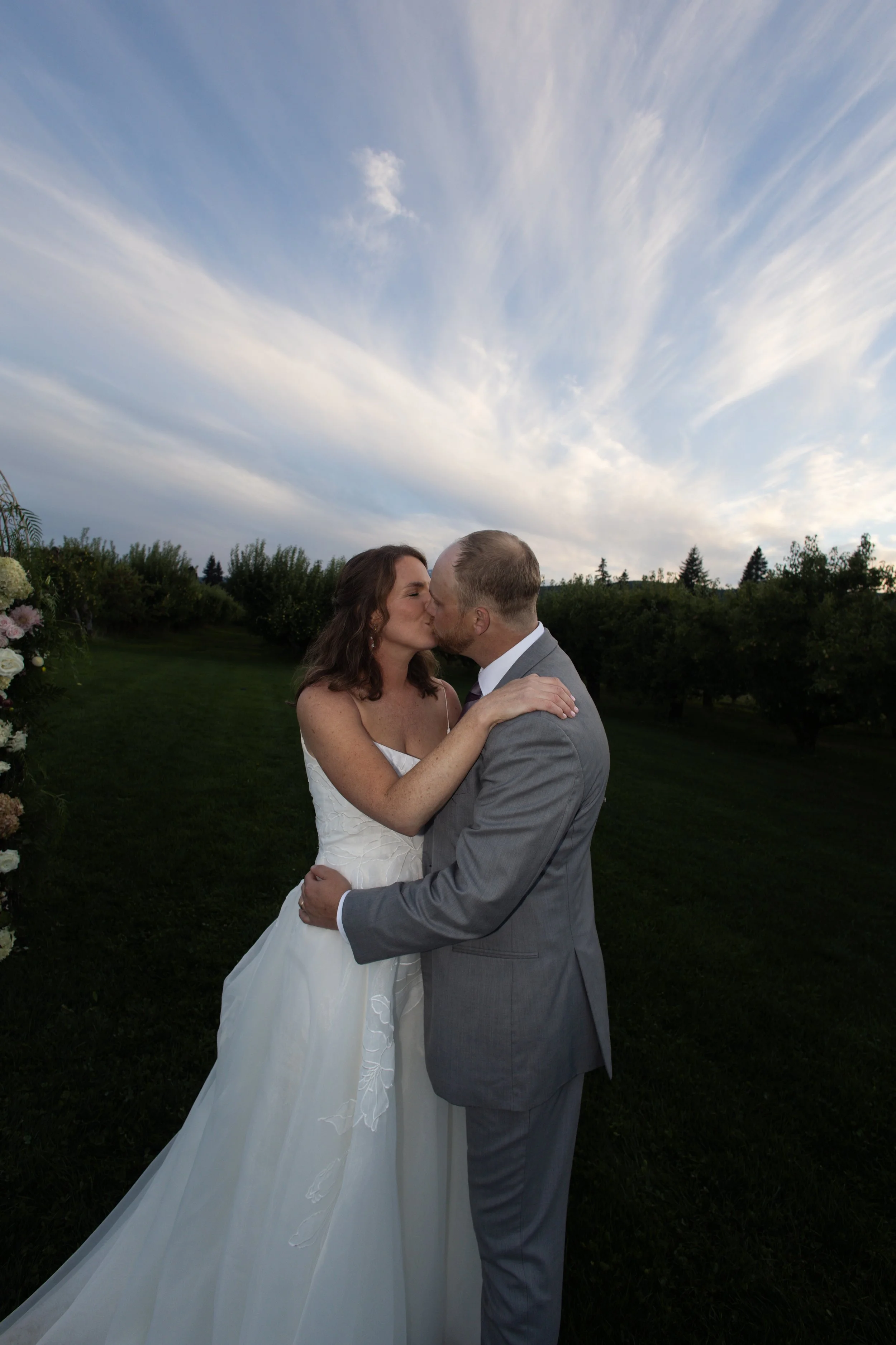 A bride and groom kissing outdoors on their wedding day with a cloudy sky above and trees in the background.