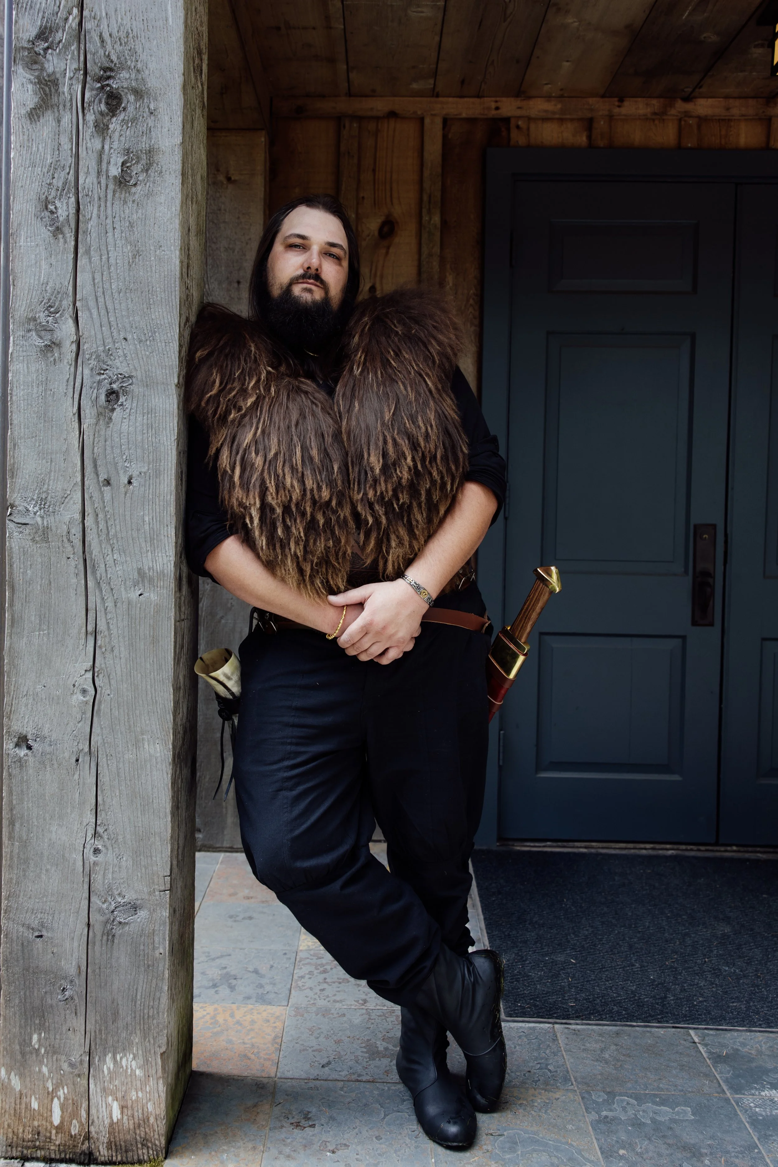 A man dressed in medieval warrior costume with a large fur cloak, holding a sword, standing outdoors near a wooden wall and black door.