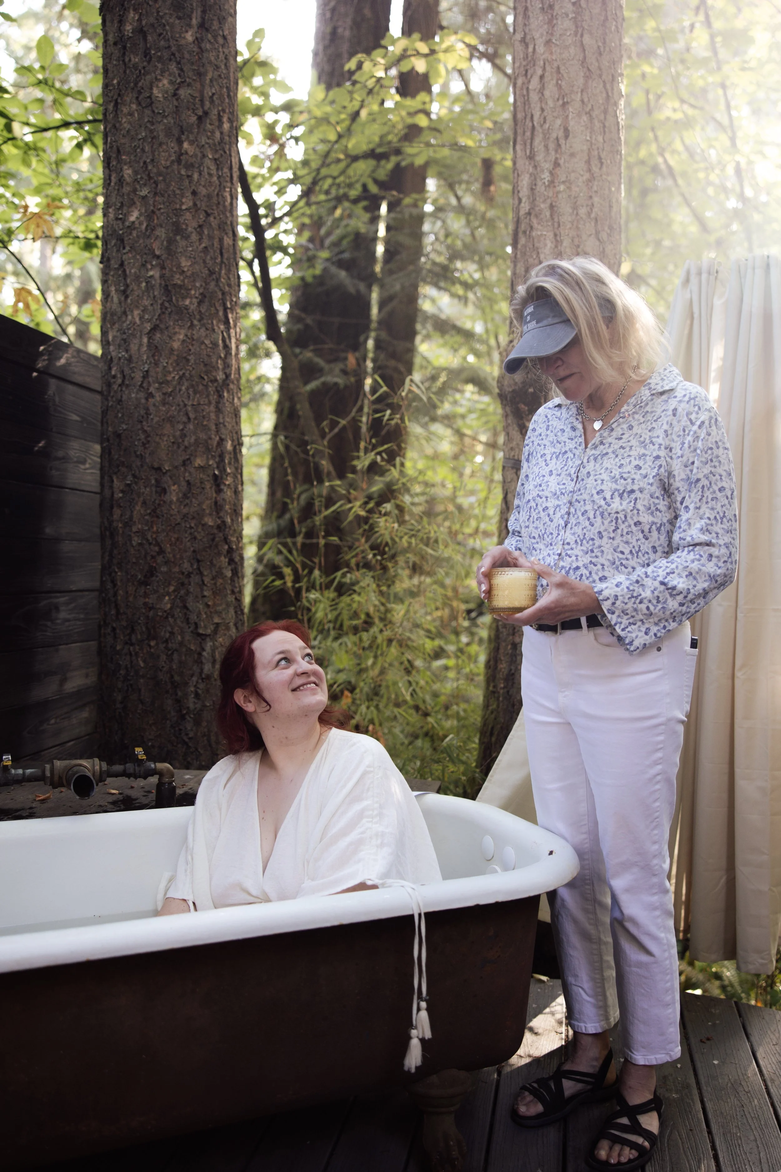 A woman with blonde hair, wearing a visor hat and floral shirt, is standing outdoors in a wooded area. She is holding a container and looking down at a woman with red hair, who is sitting in an old bathtub, wearing a white shirt. The scene appears to