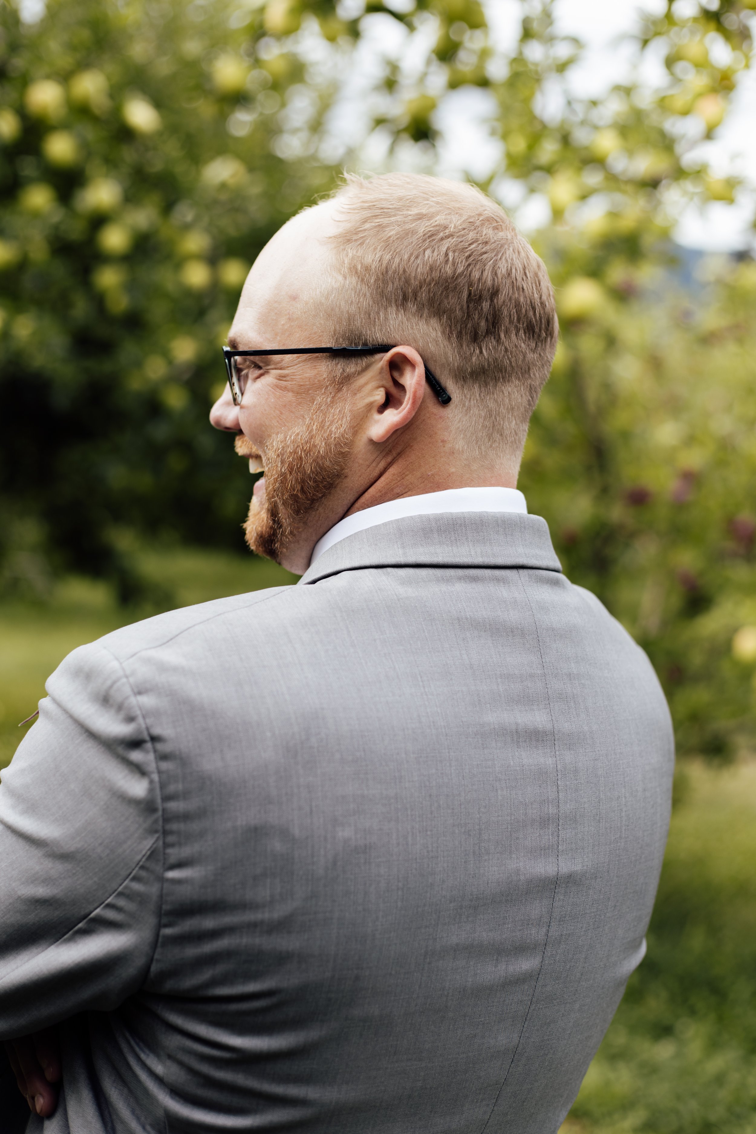 Side view of a man with a beard, wearing glasses and a light gray suit, outdoors with green trees in the background.