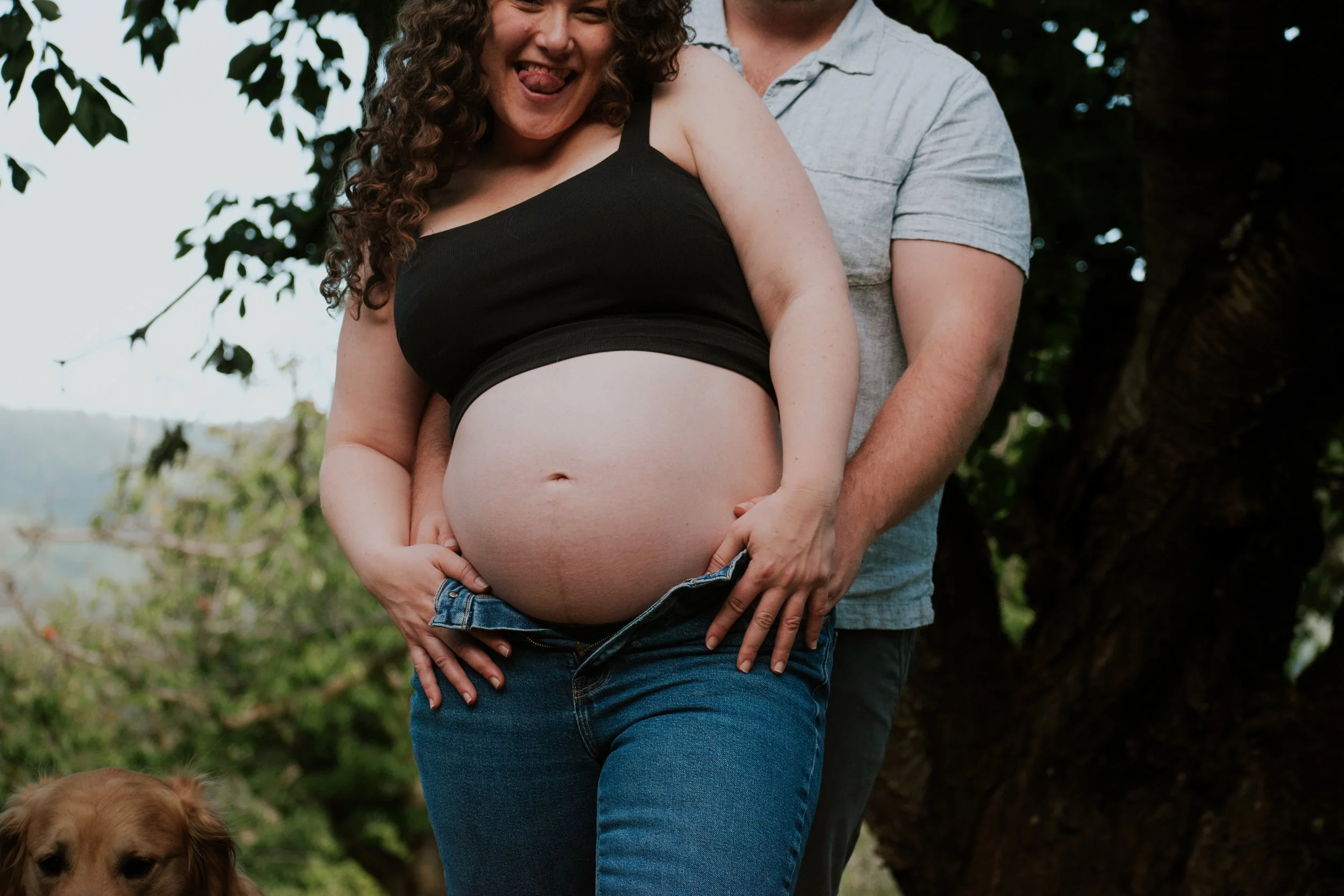 A pregnant woman with curly hair sticking out her tongue, wearing a black crop top and blue jeans, stands outdoors with a man partially visible behind her, next to a tree. The man is wearing a light-colored shirt. There is a golden retriever dog at t