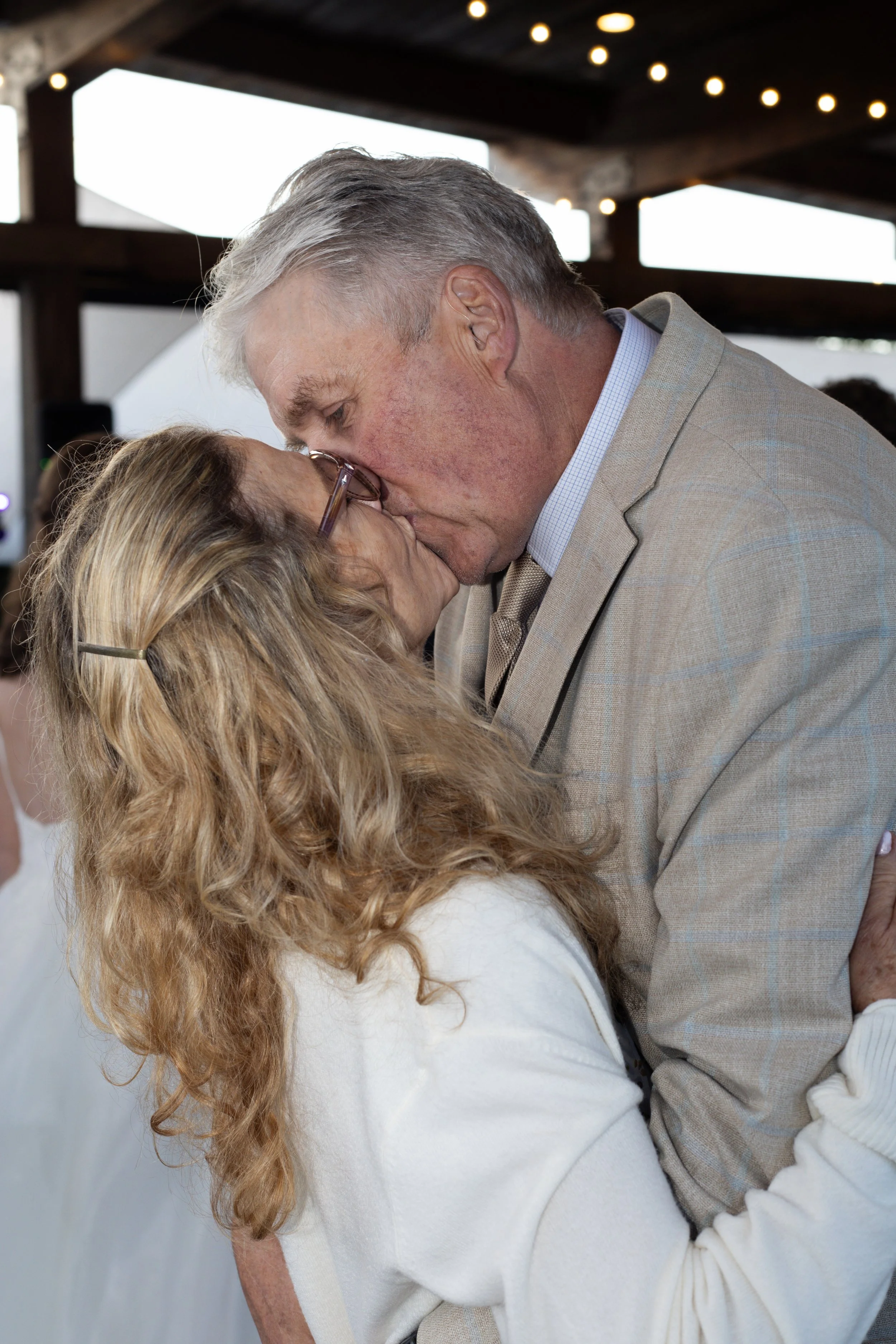 A man and a woman share a gentle kiss inside a rustic venue with string lights overhead.