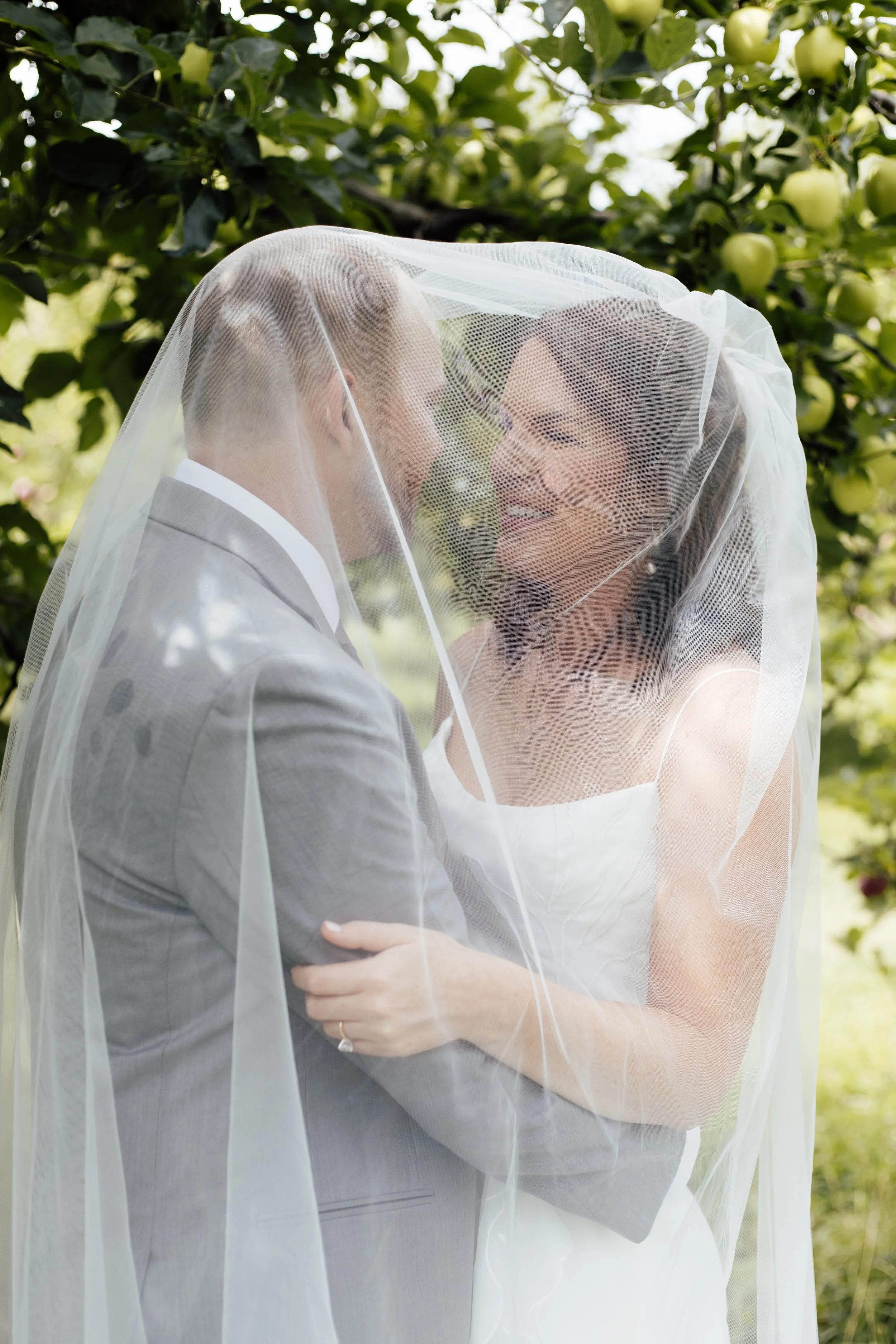 A bride and groom facing each other under a veil, smiling, outdoors with green apple trees in the background.