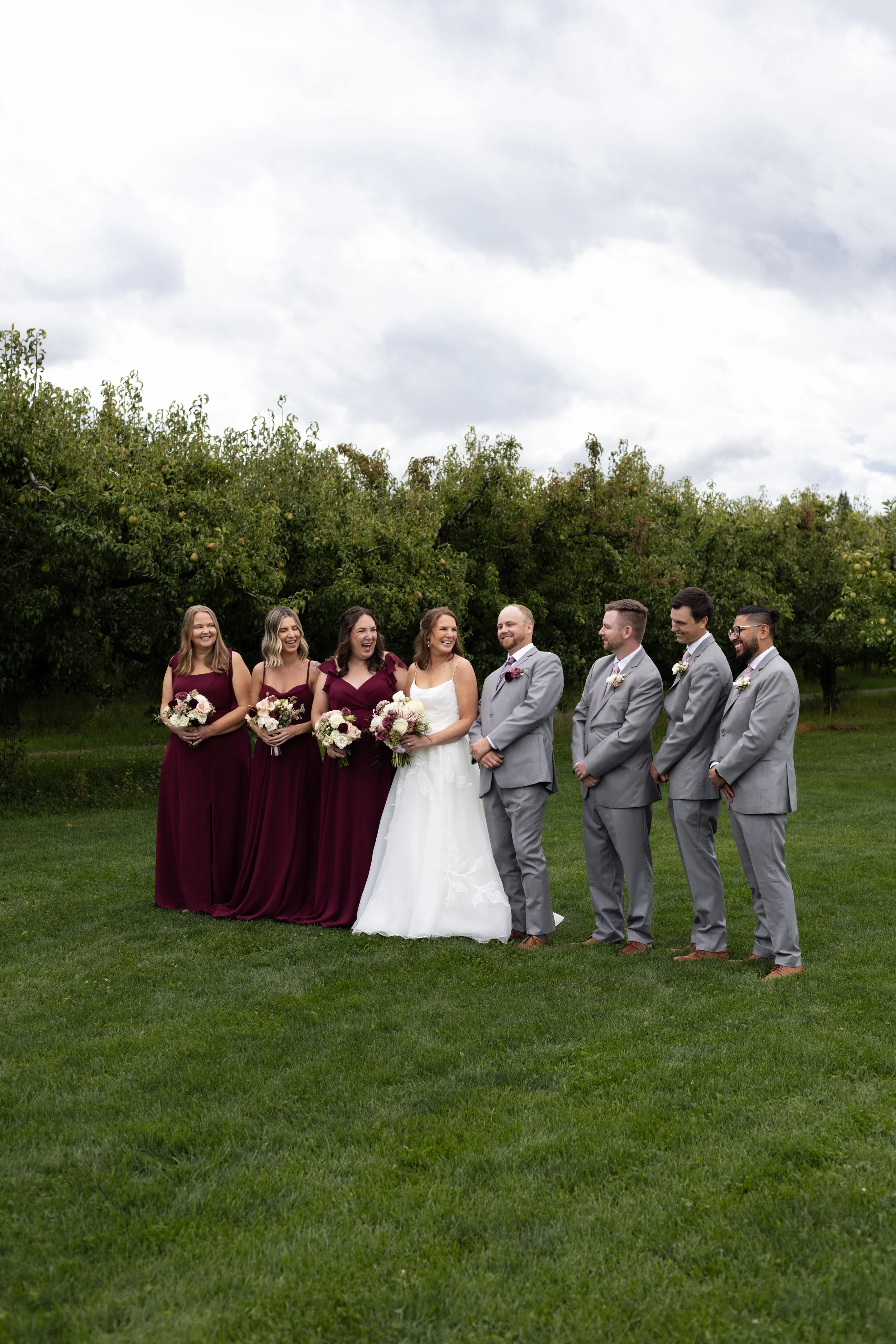A wedding party posed outdoors on a grassy field with trees and cloudy sky in the background. The bride and groom are in the center, with the bride in a white dress and the groom in a gray suit. Bridesmaids in burgundy dresses and groomsmen in gray s