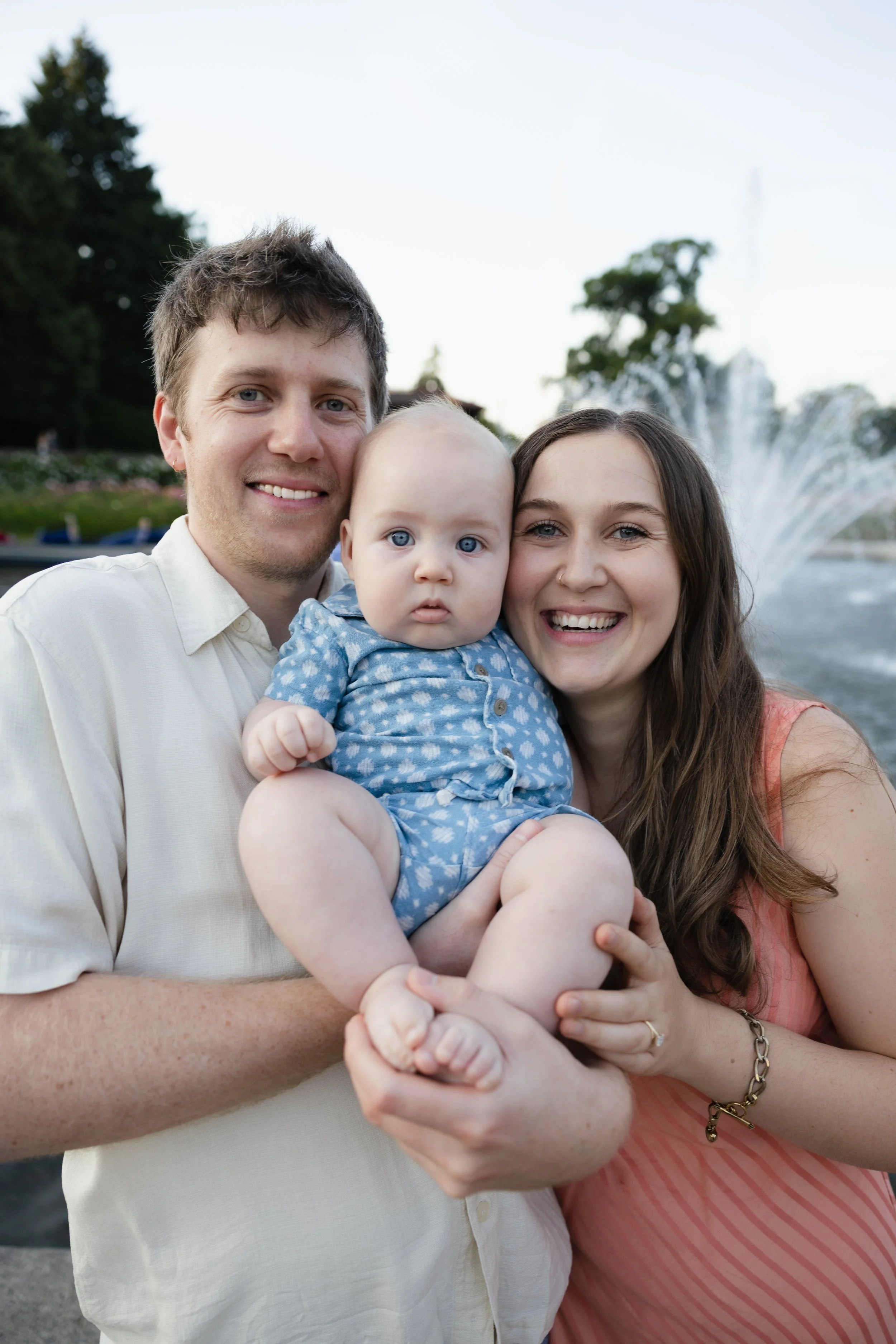 A smiling family with a man, woman, and baby posing outdoors near a fountain and trees.