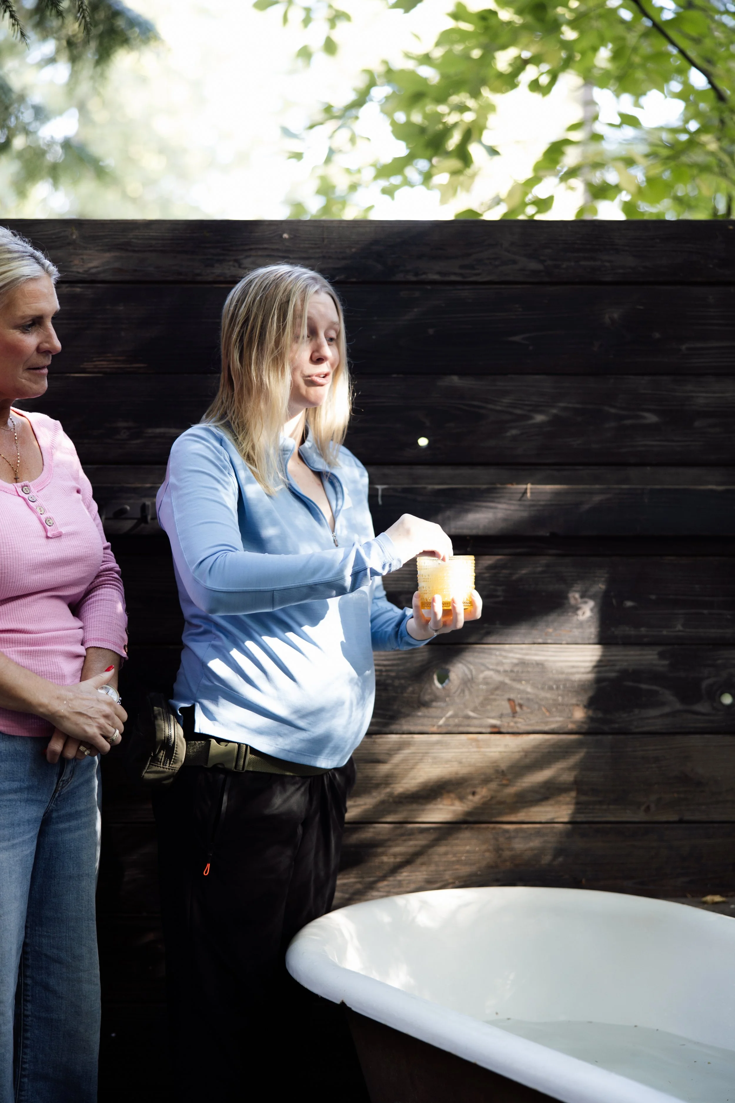 Two women standing outdoors near a wooden fence, one holding a candle, with a white bathtub in the foreground.