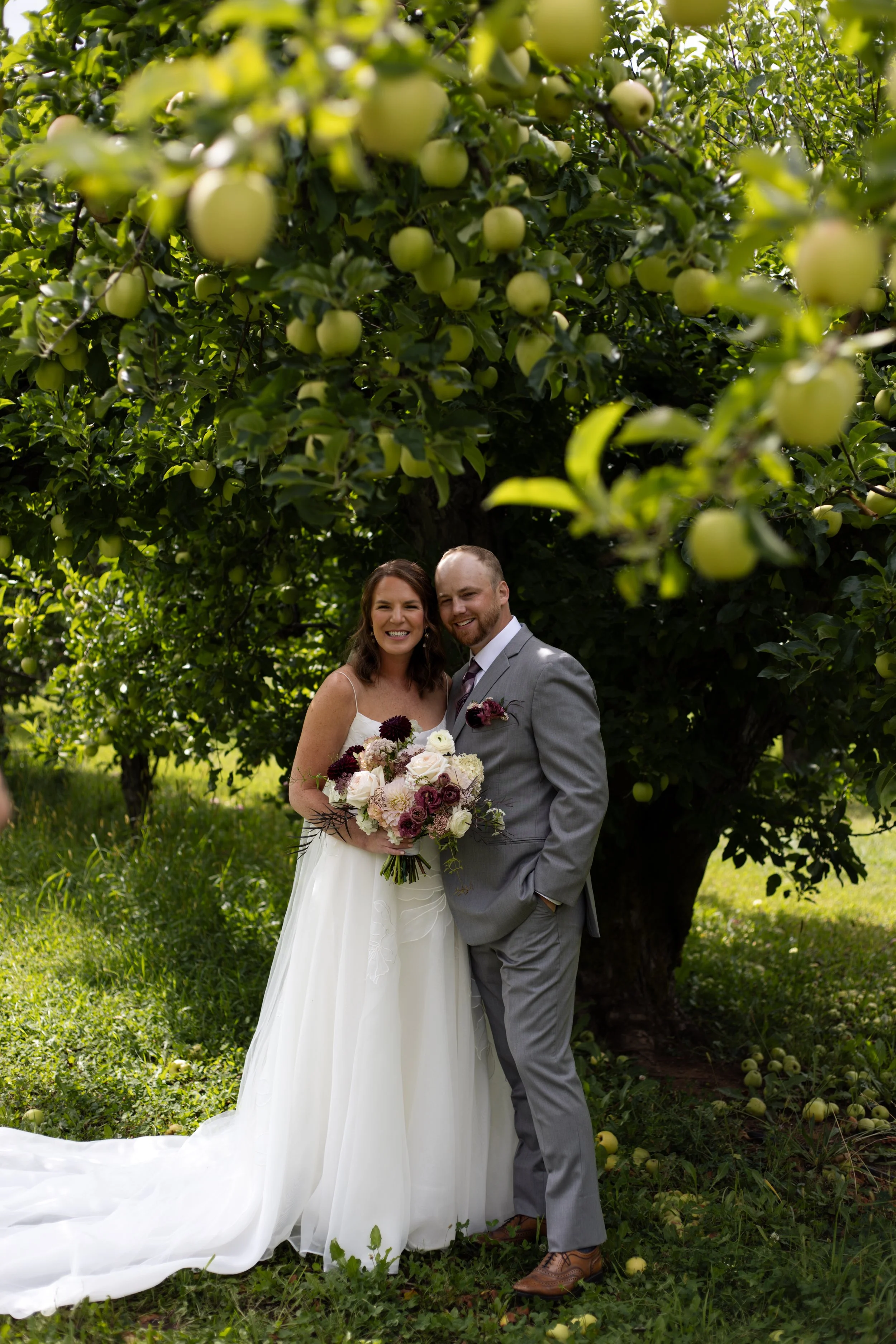 A bride and groom standing together outdoors in front of a large apple tree, smiling and holding a bouquet of flowers, with green grass and fallen apples on the ground.