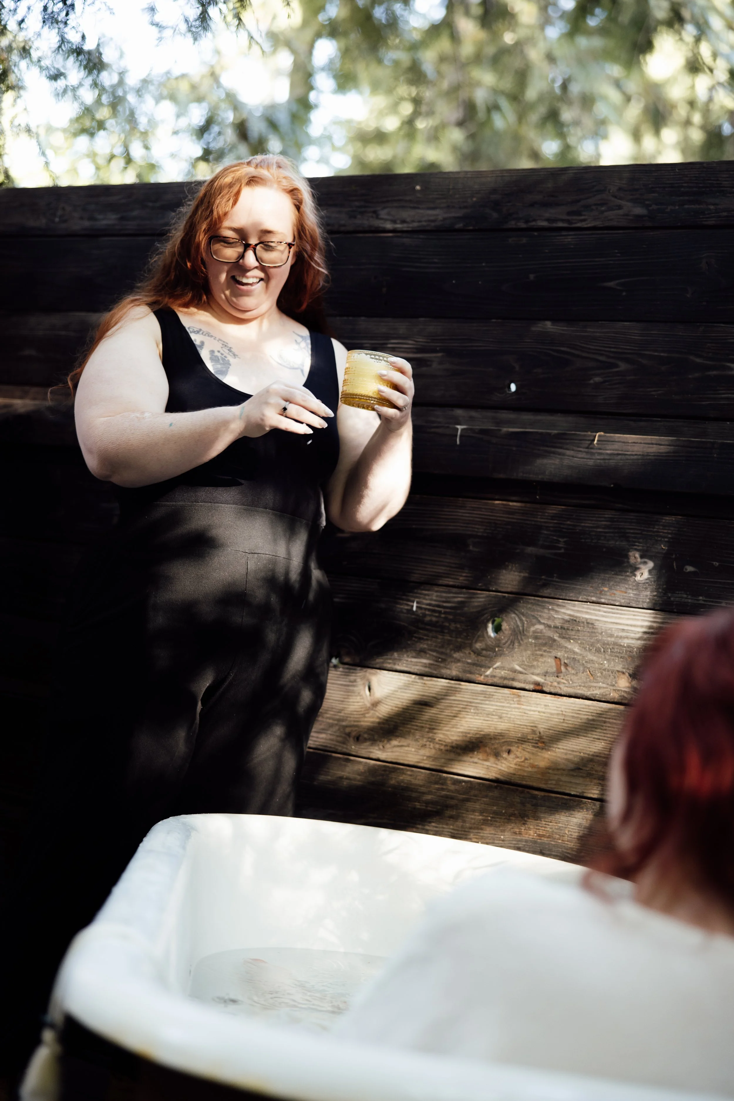 A woman with red hair and glasses smiling while holding a drink, standing outside in front of a wooden fence with trees in the background.