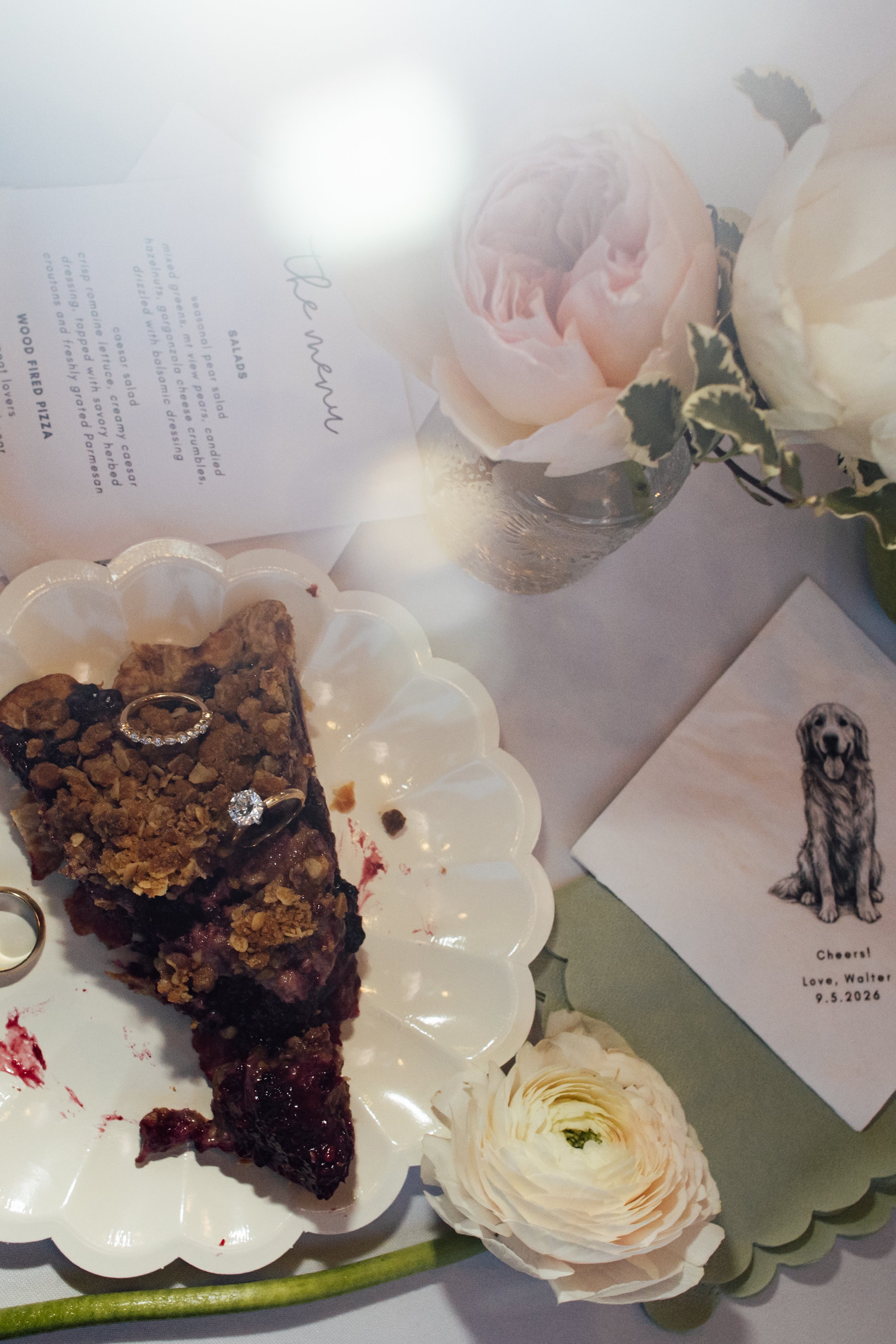 A slice of cherry dessert topped with crushed nuts on a white scalloped plate, decorated with white roses and greenery, with a background of printed thank you notes and a dog illustration.