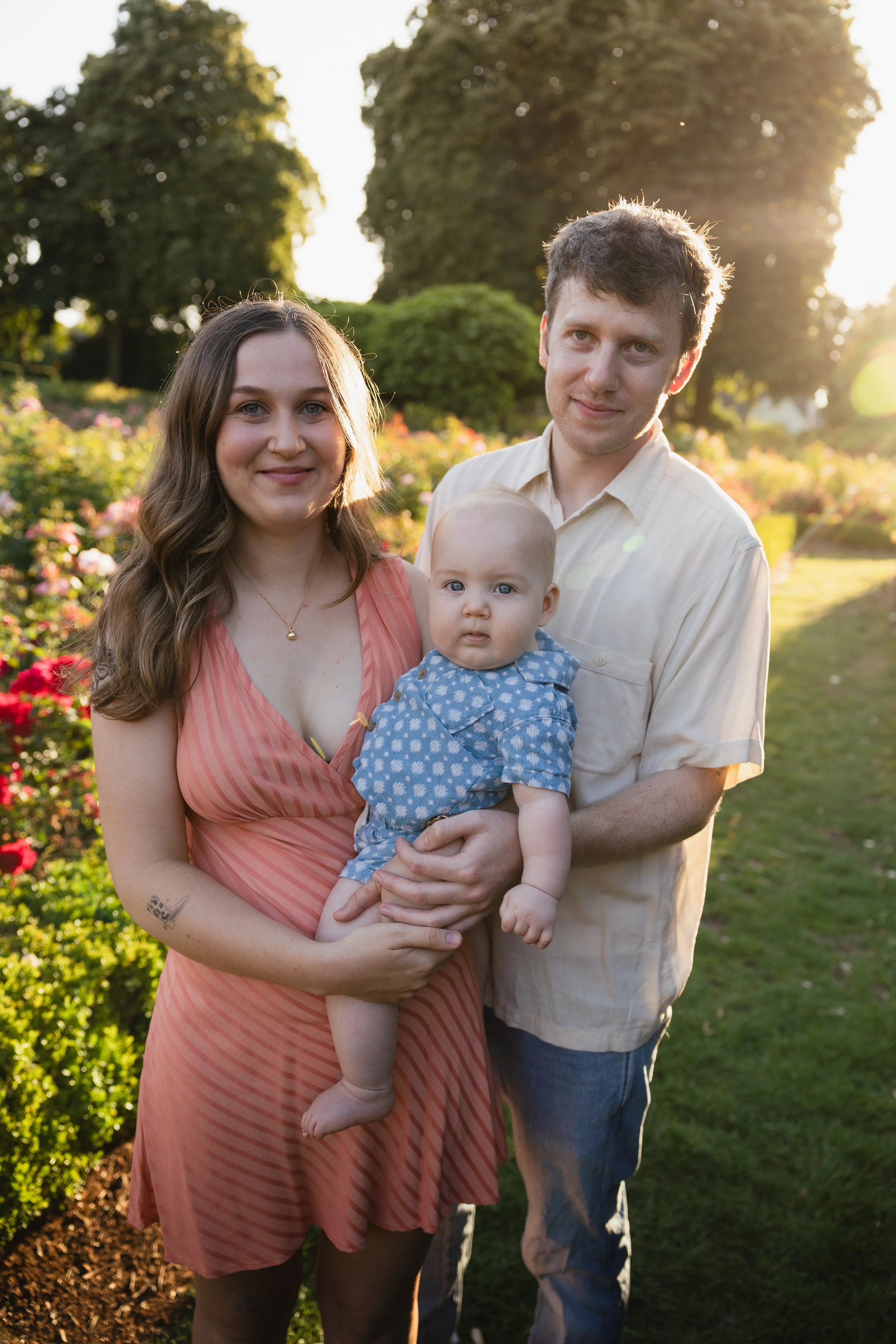 A family of three standing outside in a garden during sunset, with trees and blooming flowers in the background.