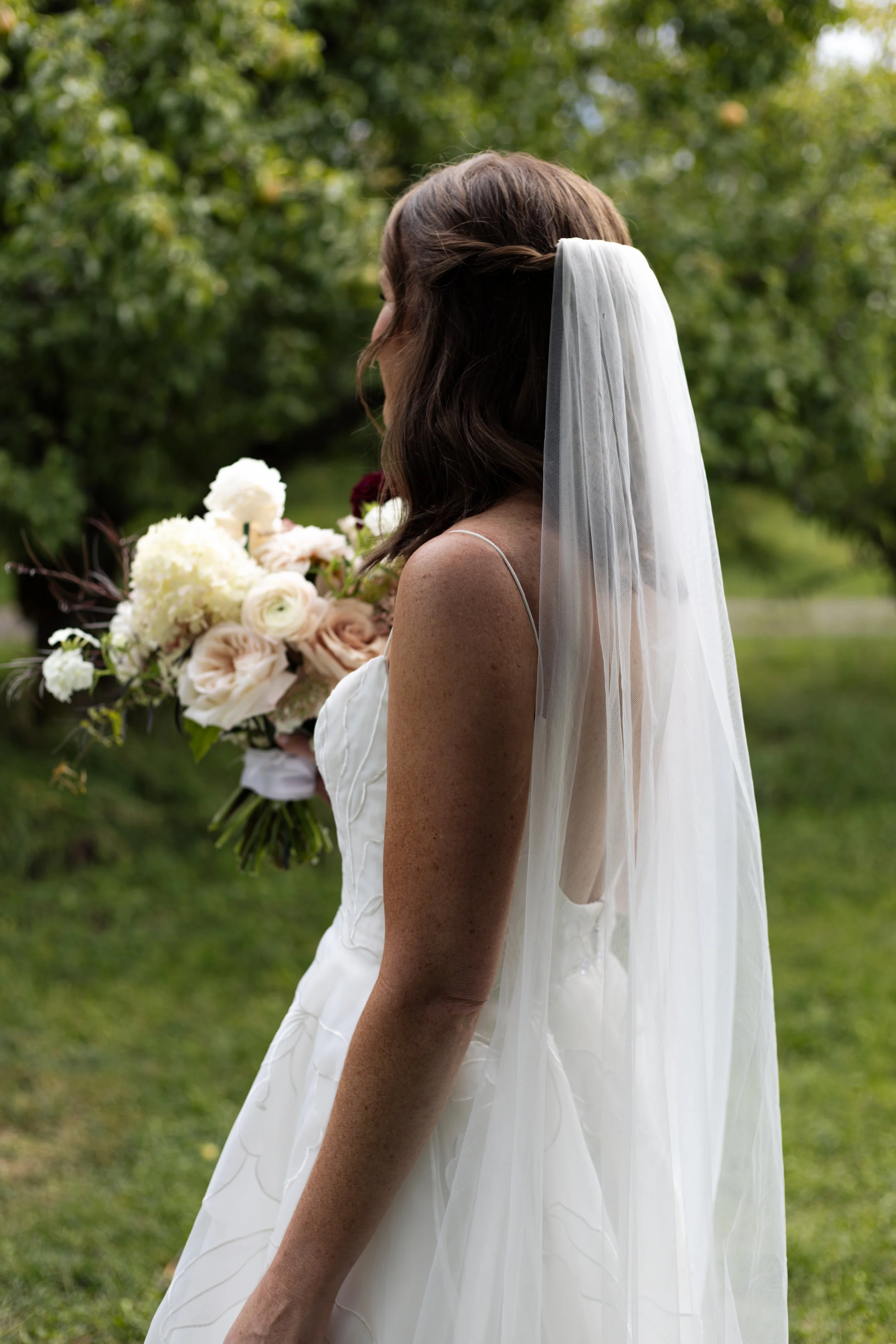 A woman in a wedding dress with a veil holding a bouquet of white, pink, and dark flowers outdoors in a green park.