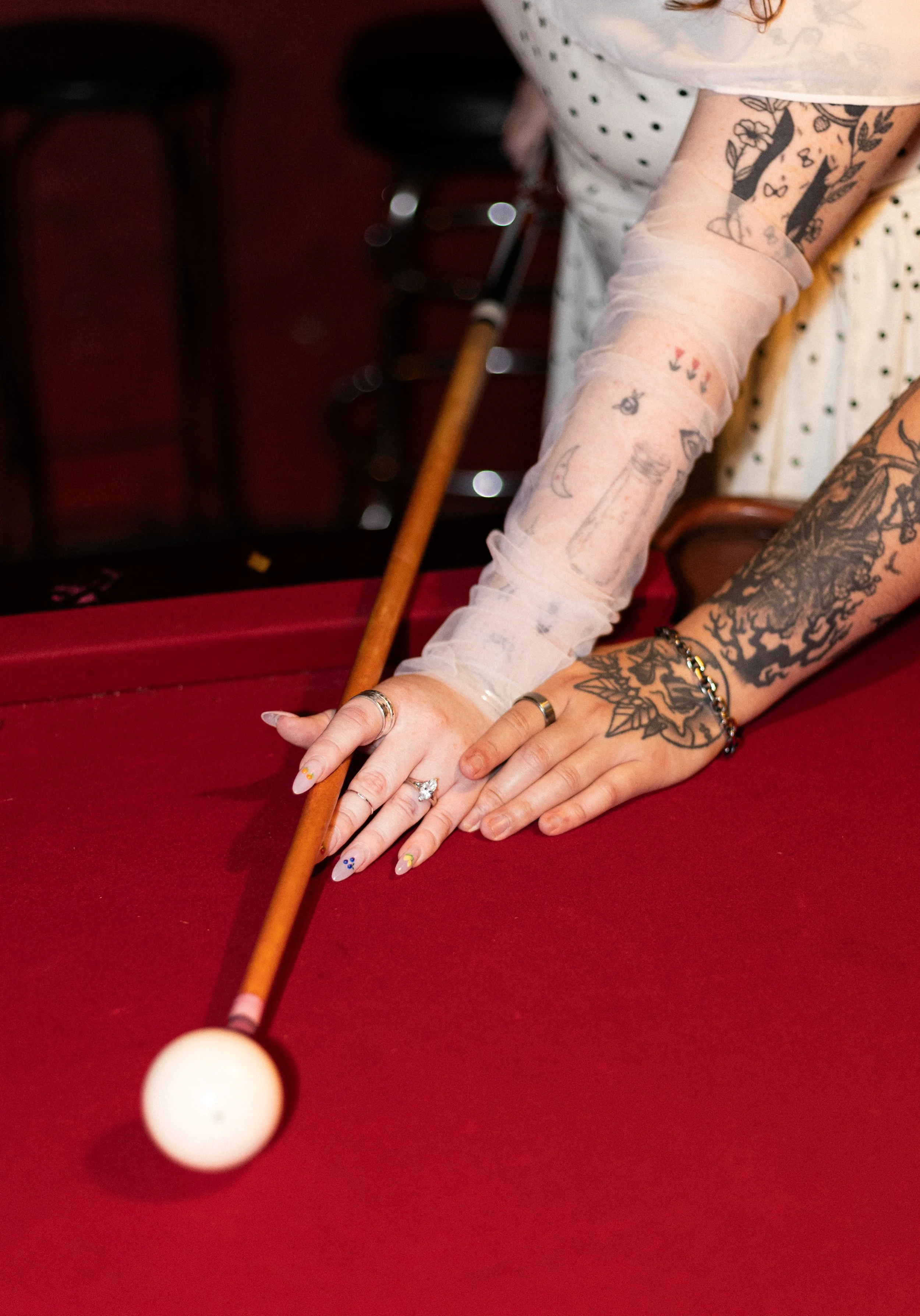 Close-up of a person playing pool, with tattoos on their arms and colorful nails, aiming at the cue ball on a red felt pool table.