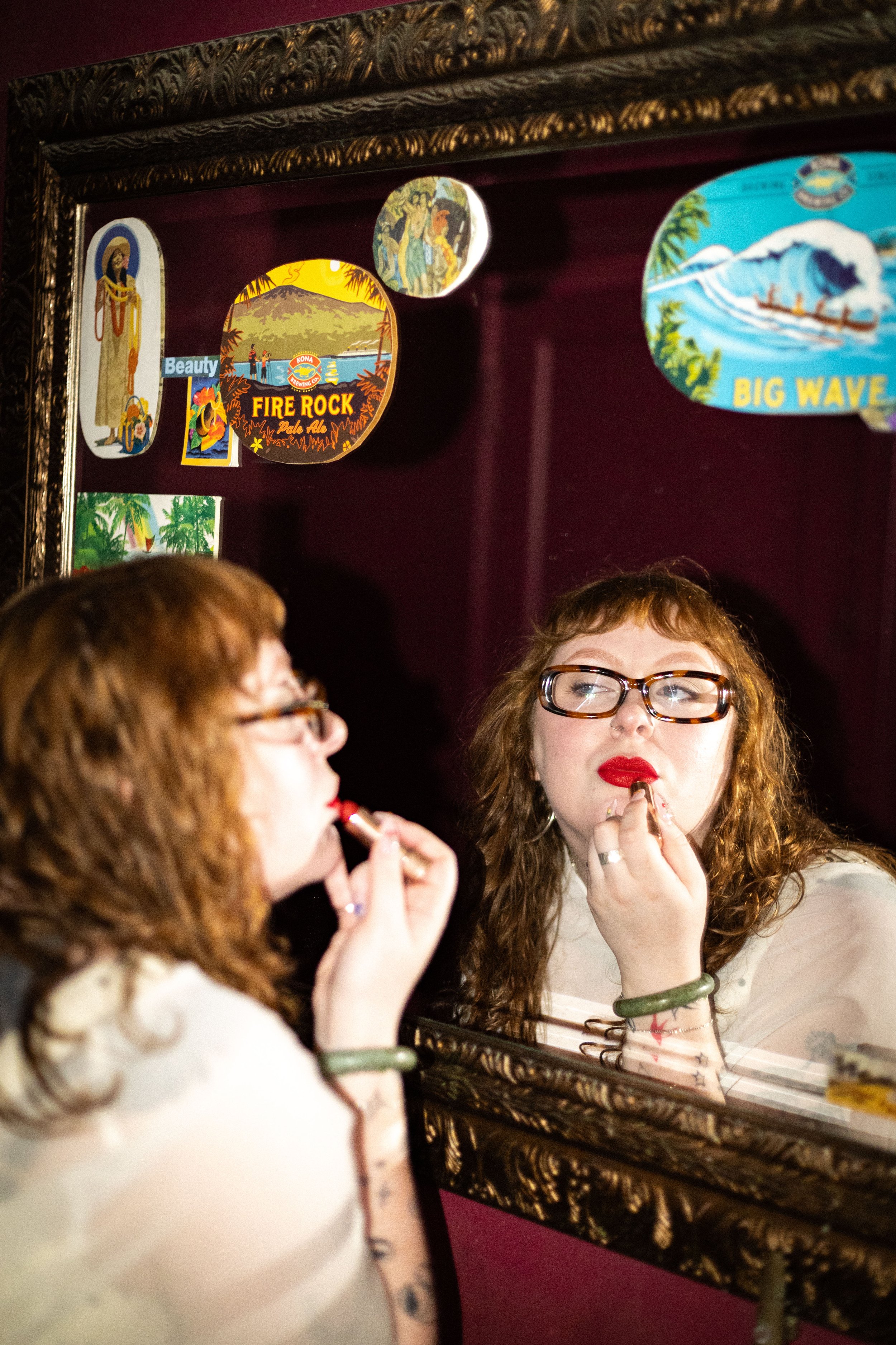 A woman with curly hair and glasses applying lipstick while looking into a mirror, with a portrait and decorative items on a burgundy wall in the background.