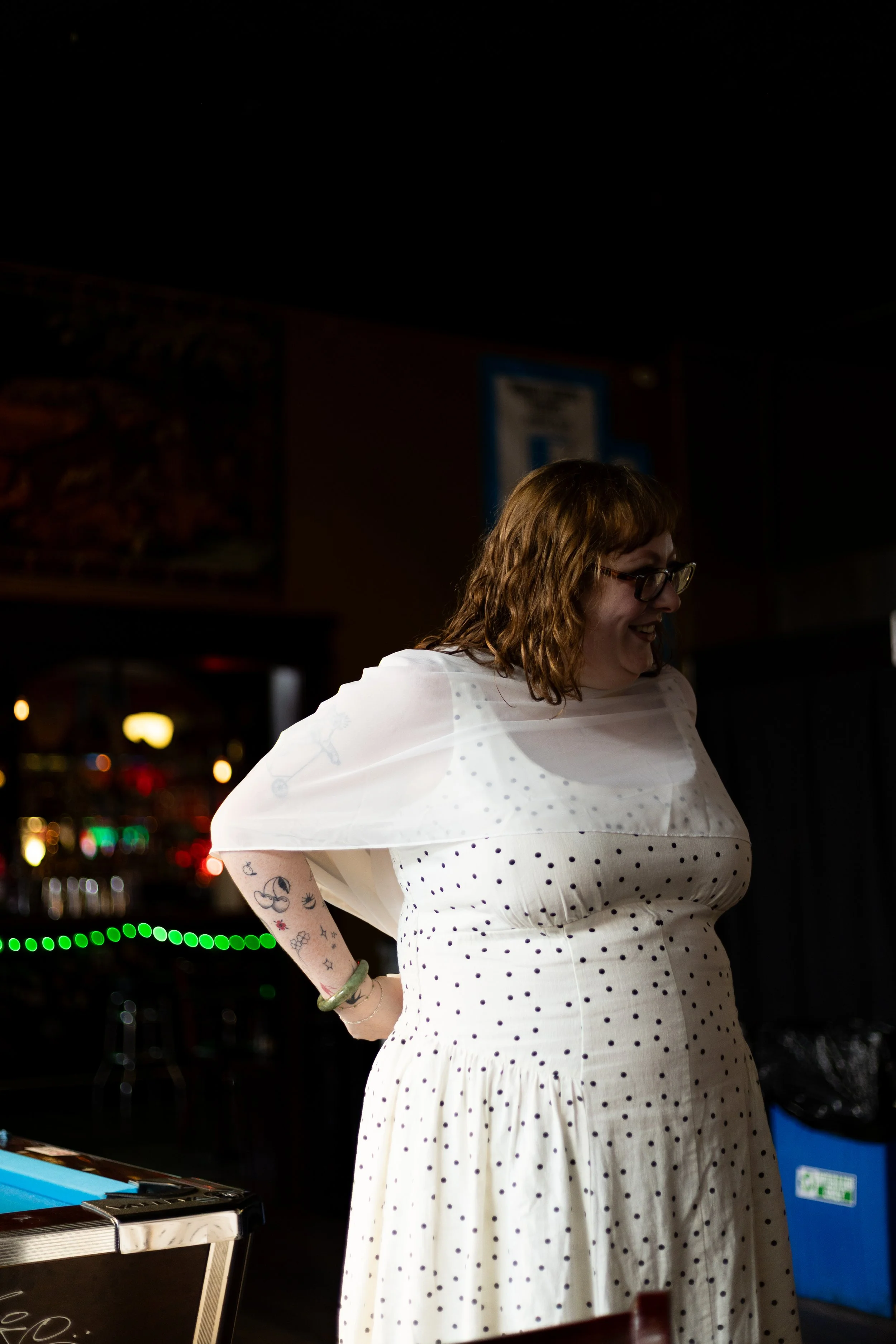 A woman with curly brown hair and glasses wearing a white dress with black polka dots, smiling in a dimly lit restaurant or bar with drinks and signs in the background.