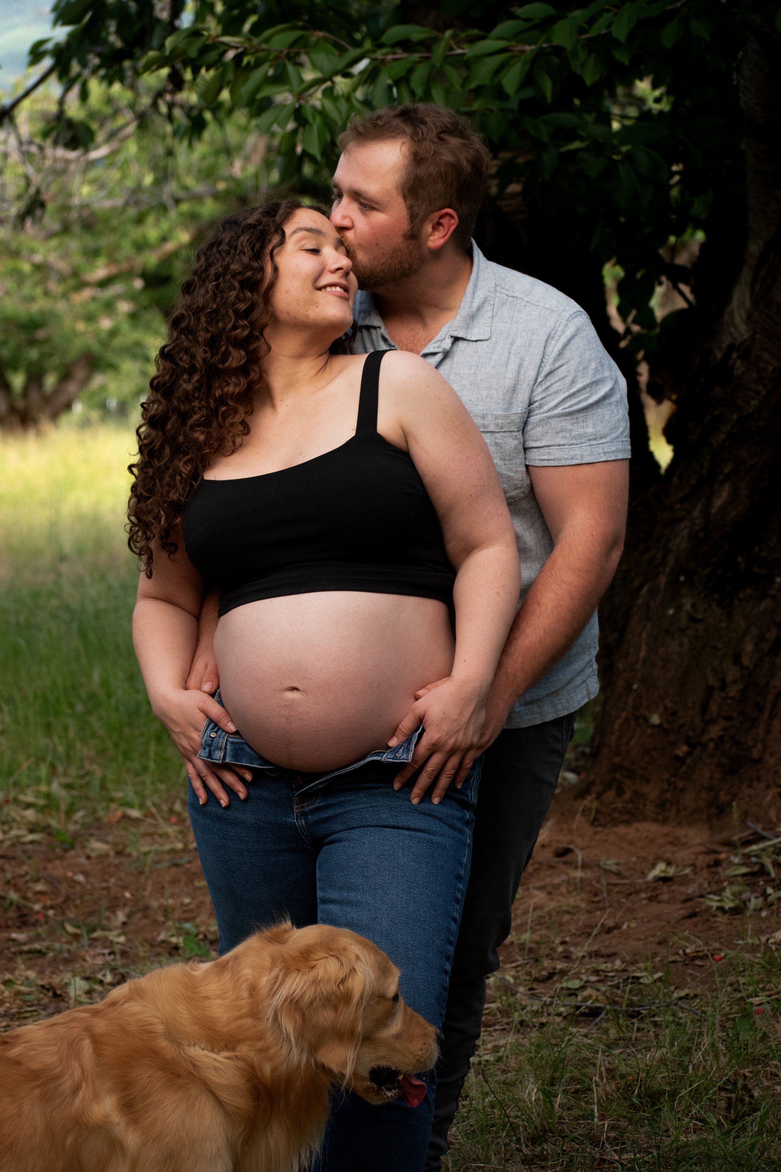 A pregnant woman with curly hair in a black tank top and jeans, standing outdoors near a man behind her, who is kissing her forehead. The man has short hair and a beard, wearing a light gray shirt. A large golden retriever is in the foreground, sitti