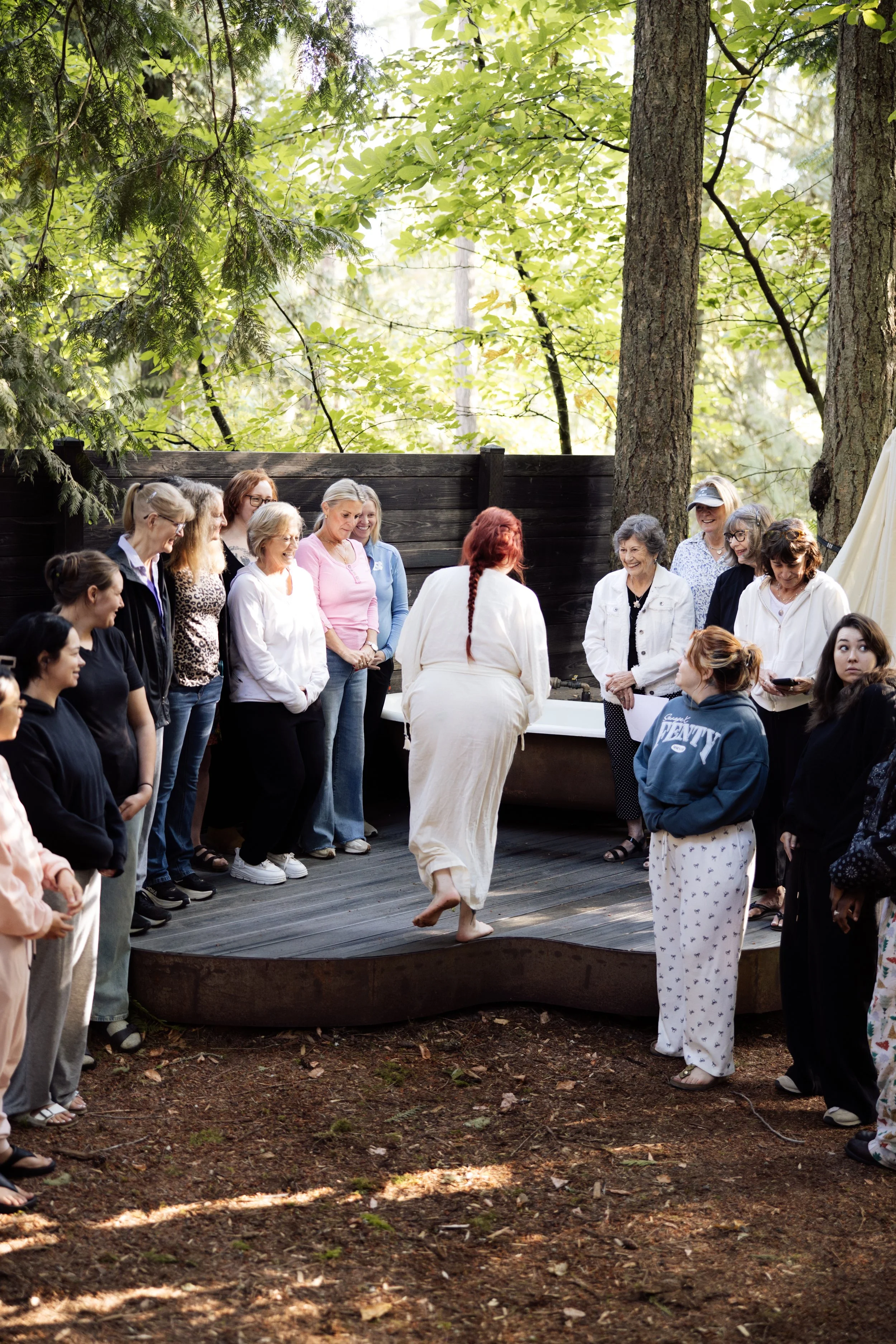 A group of women gathered outdoors around a woman in white dancing on a stage, with trees and green foliage in the background.