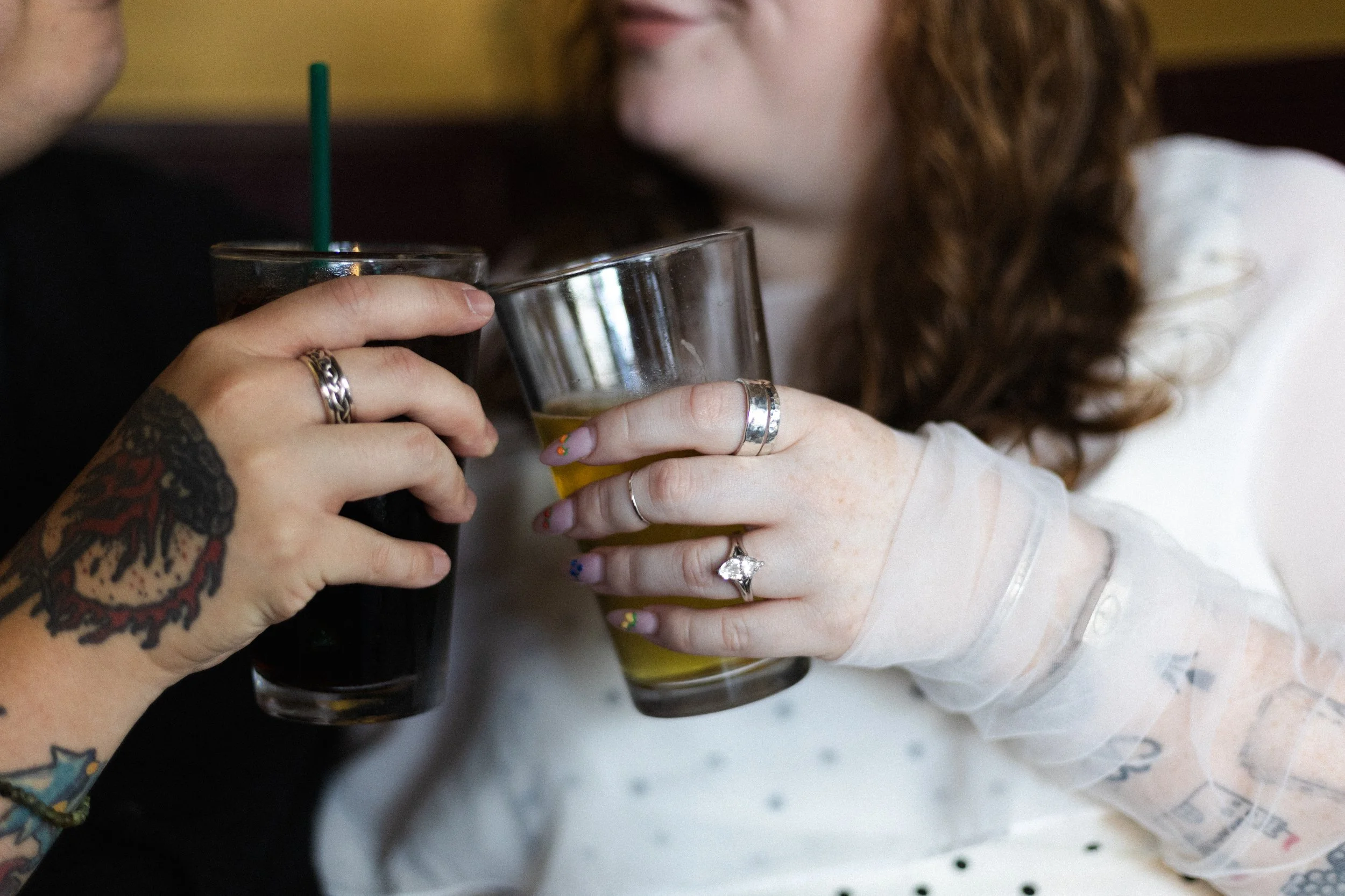Two people holding glasses of dark soda and beer, with one person wearing multiple rings and having tattoos on their arm, and the other person wearing rings and a white sleeve, and a woman with curly hair and a white shirt.