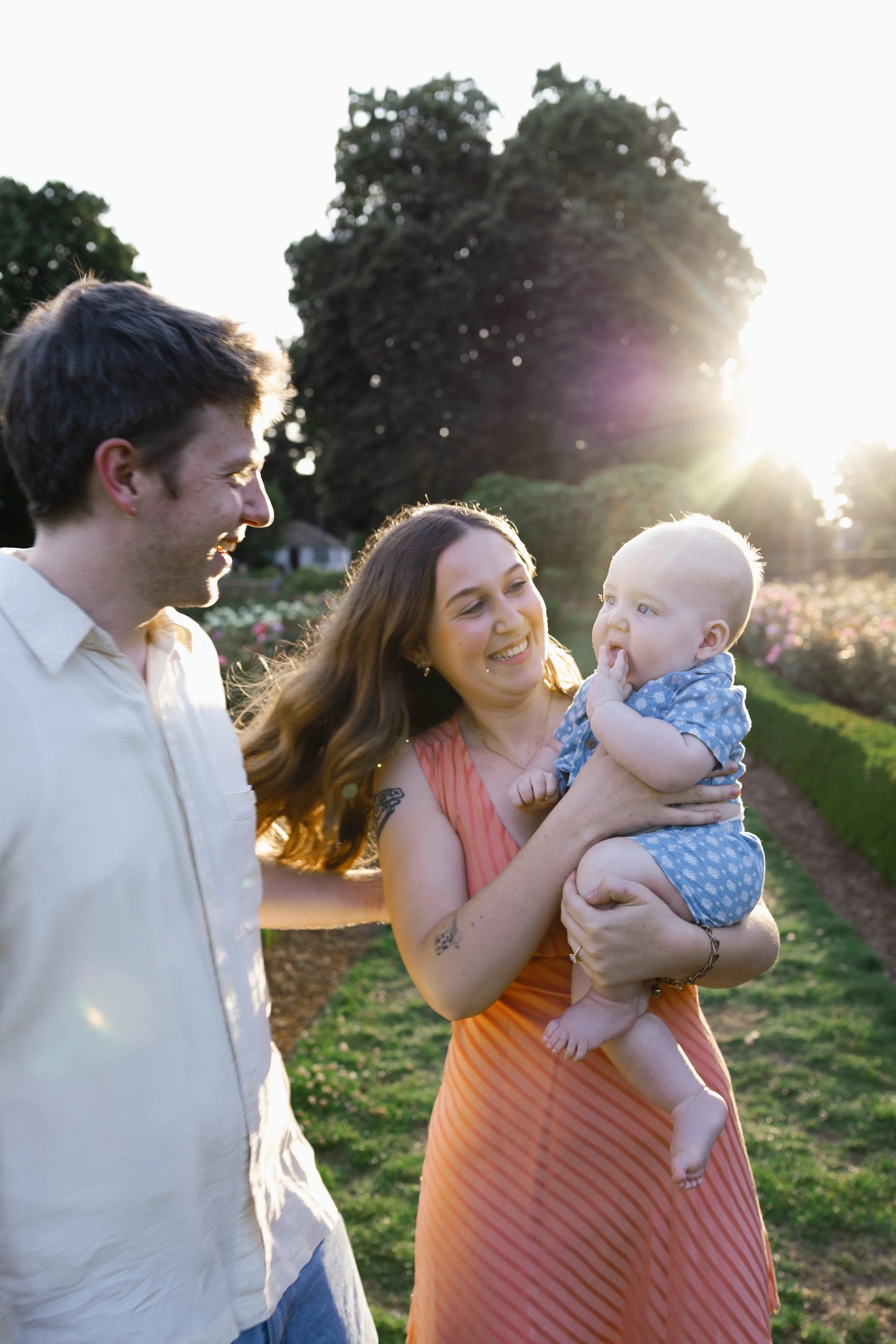A family of three enjoying time outdoors in the late afternoon or early evening sunlight, with two adults and a baby, standing in a garden or park with trees and flowers in the background.