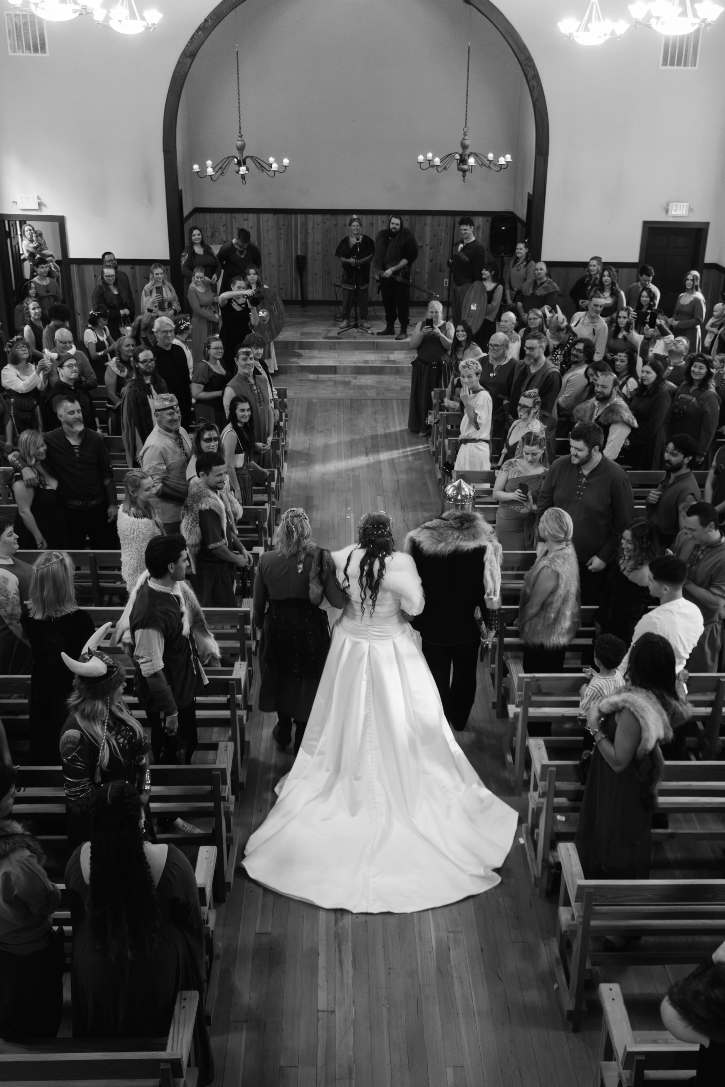 A large wedding ceremony in a church with a bride in a long white gown walking down the aisle, surrounded by guests in medieval and fantasy costumes, some with horned helmets and fur cloaks. The church interior features wooden walls, chandeliers, and an arched ceiling.