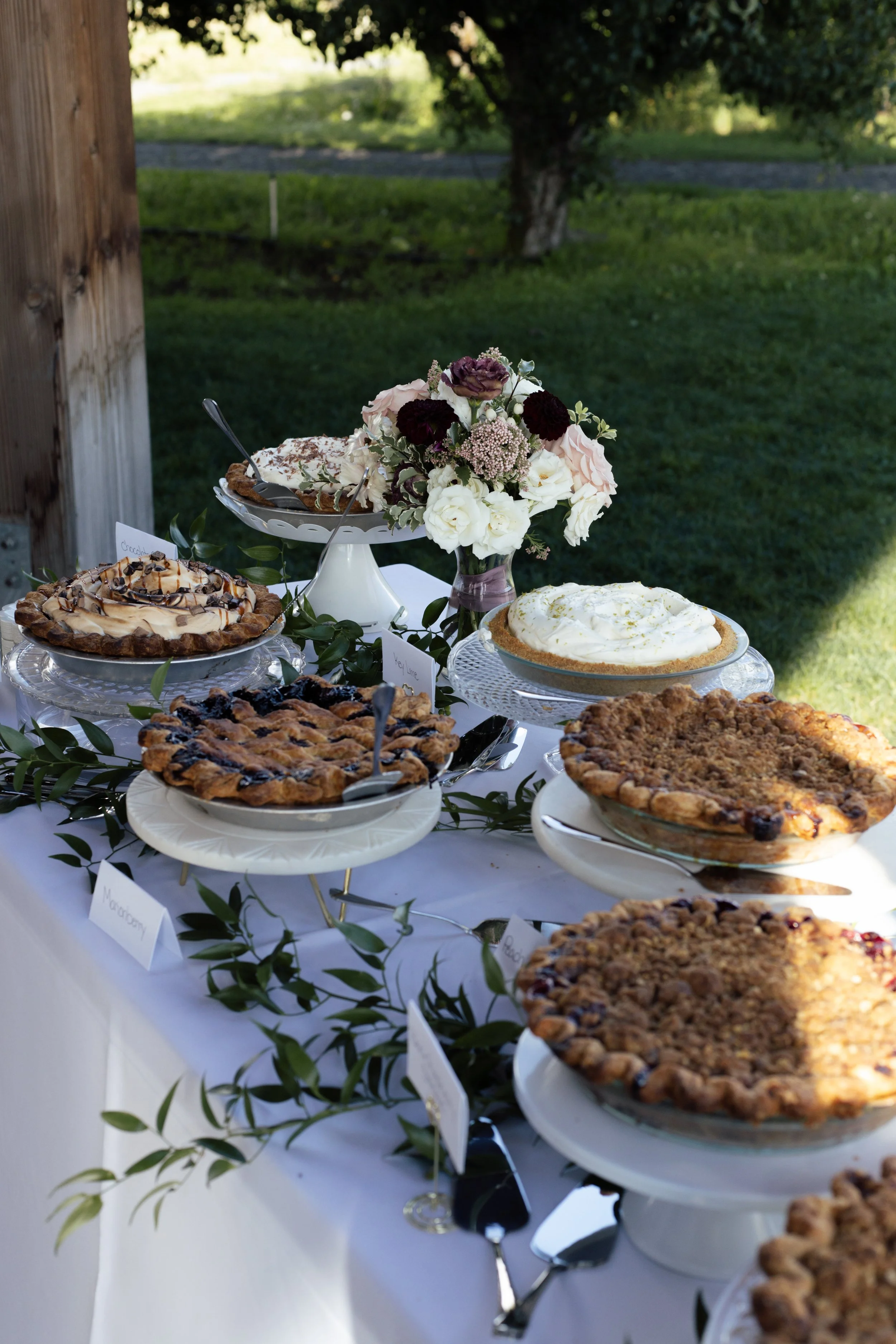 A table outdoors with assorted pies and desserts, a bouquet of white and dark purple flowers, and small labels for each dessert, set against a background of grass, trees, and a wooden post.